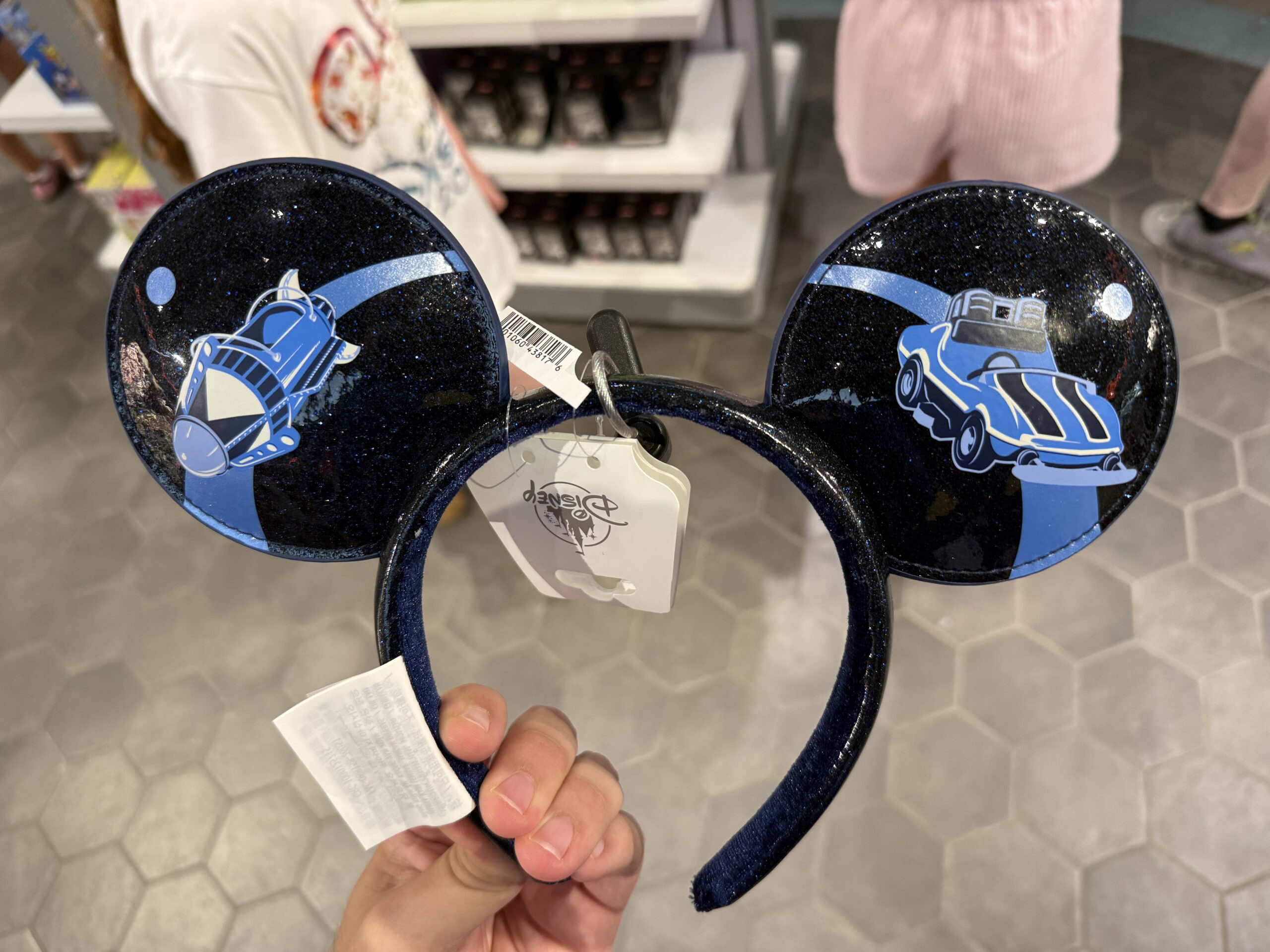 This ear headband is black and has a subtle sparkly sheen. The Space Mountain building and ride vehicle are depicted in blue on one side, while the ride vehicles from Astro Orbiter and Tomorrowland Speedway are on the other side.