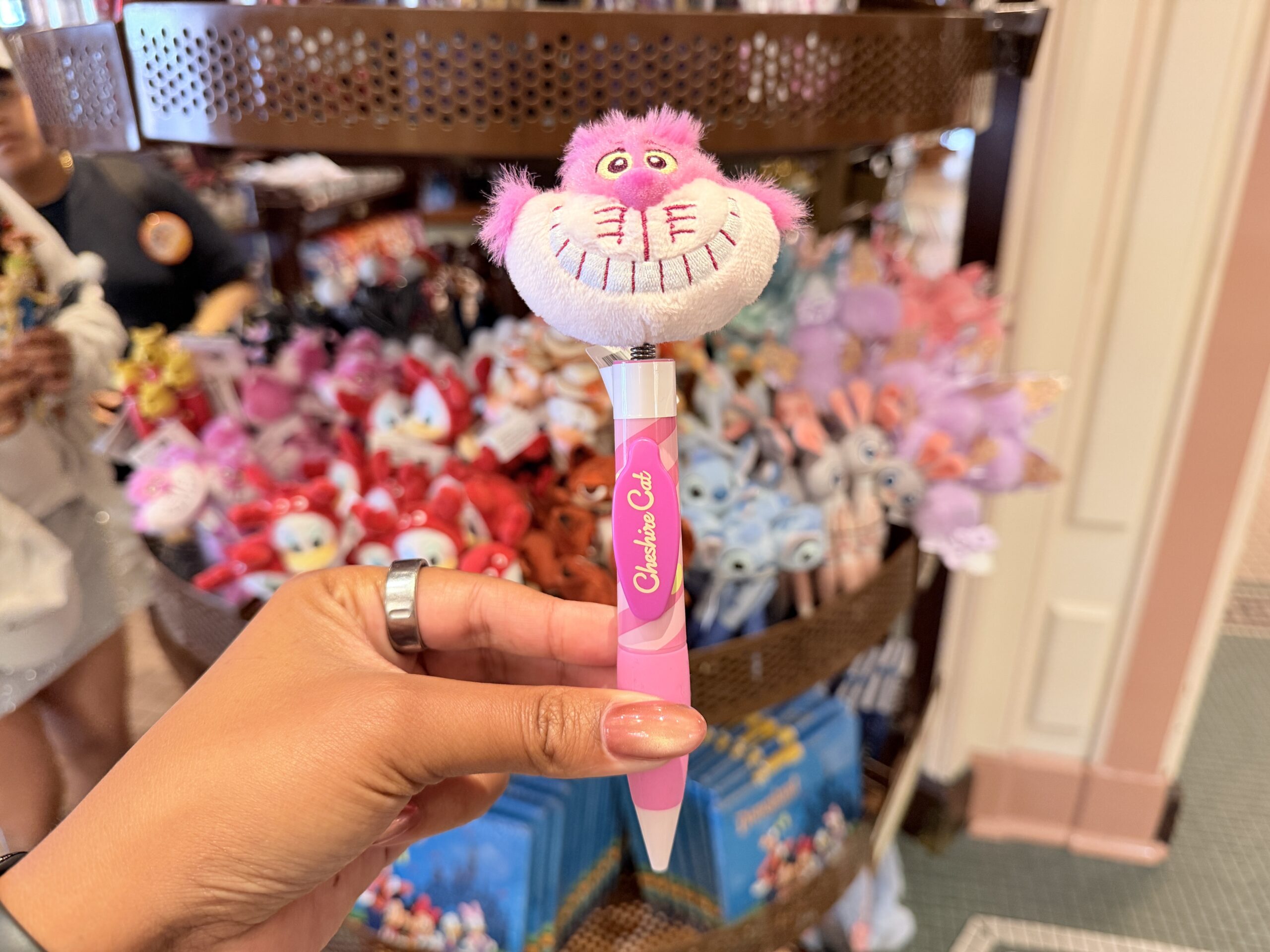 The Cheshire Cat pen features a plush pink head of the grinning Alice in Wonderland character. The topper includes his wide toothy smile, yellow eyes, and fuzzy pink fur.