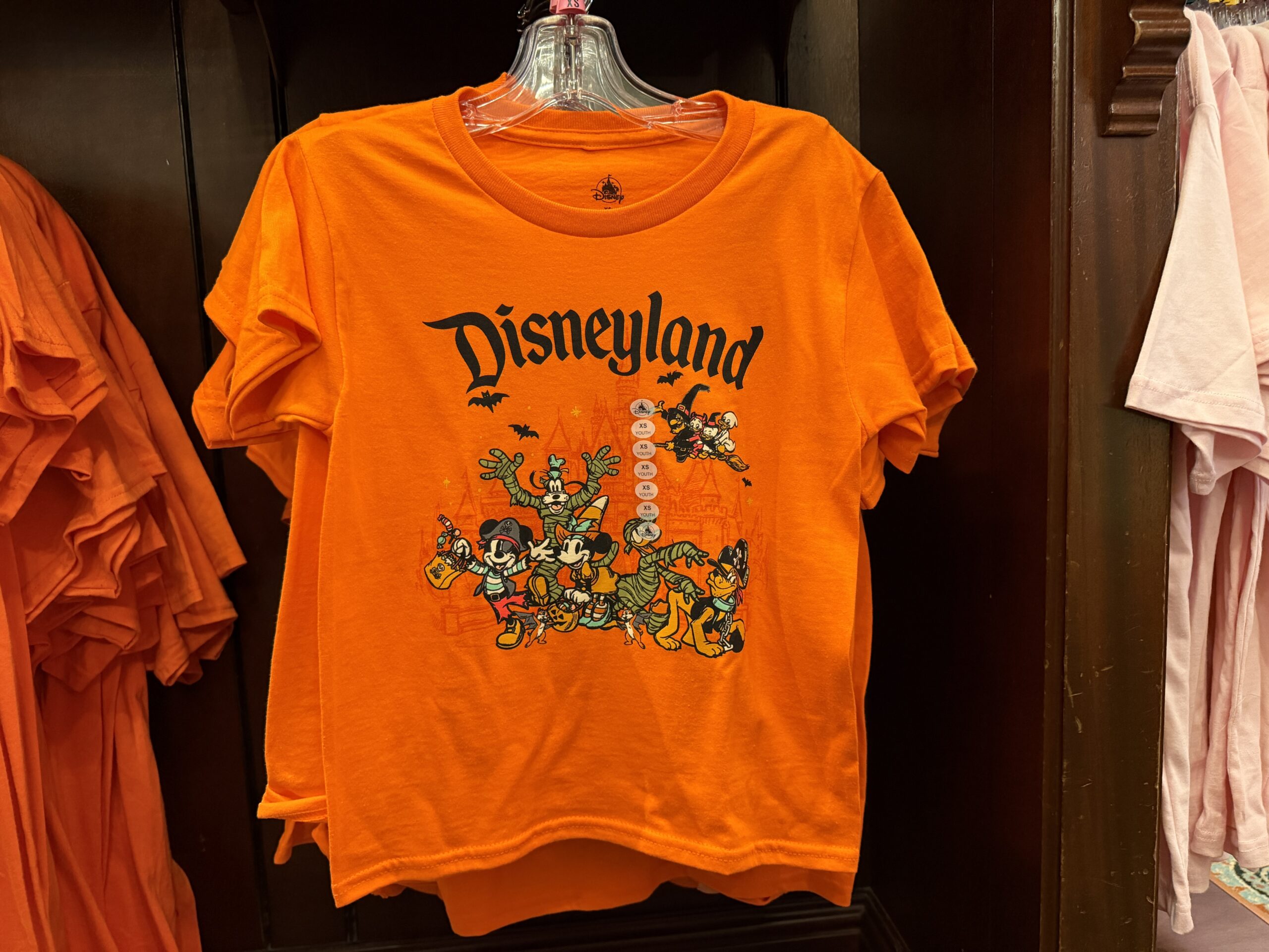 This orange tee has images of Mickey and his friends dressed in various costumes. Mickey is a pirate, Minnie is a witch, Goofy and Donald are both mummies, and Pluto is also a pirate. Behind them, bats fly and a witch carries Huey, Dewey, and Louie on a broom. The Disneyland wordmark is at the top in black.