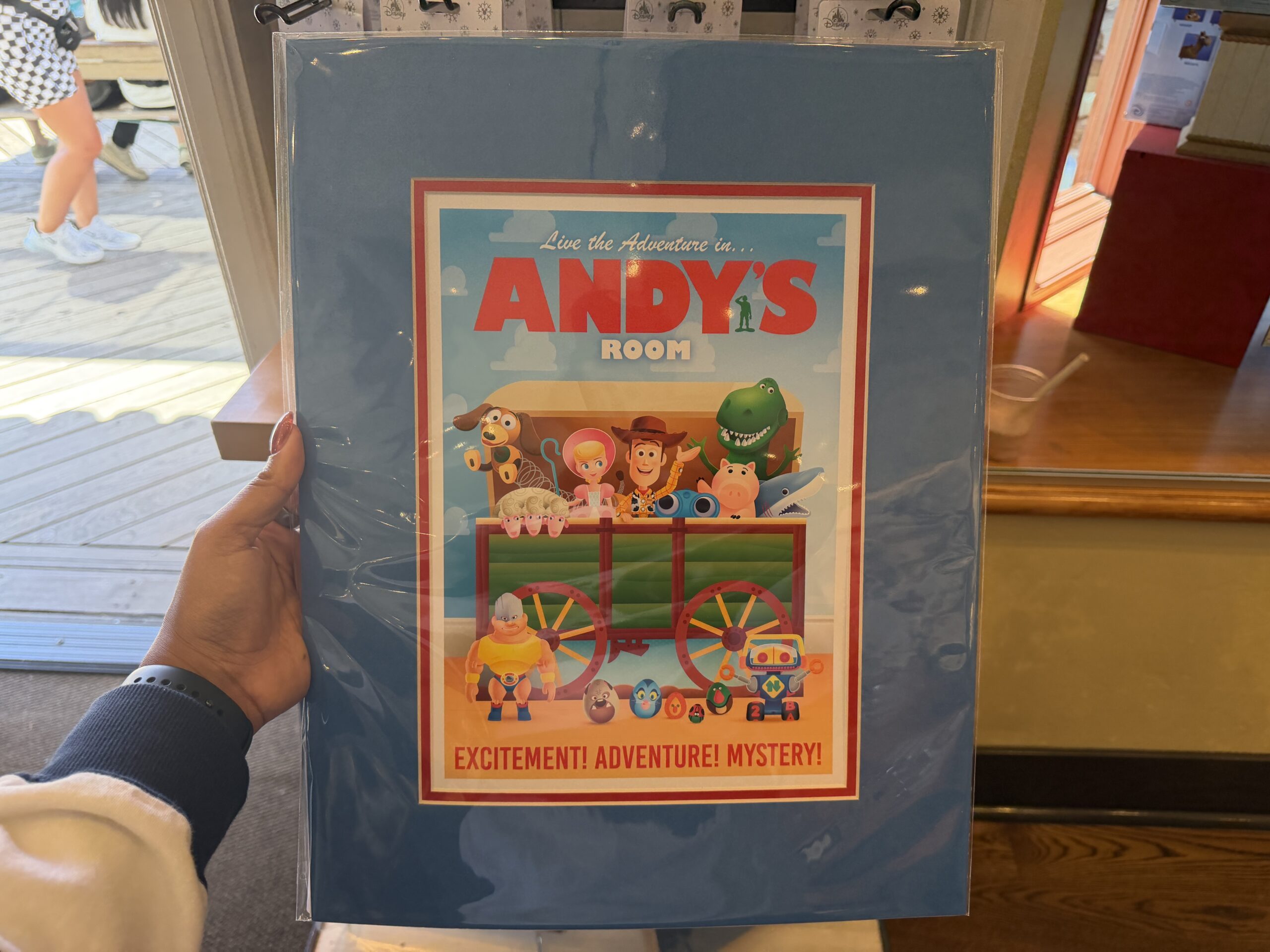 This piece in Jerrod Maruyama's style shows several of the original Toy Story characters in Andy's covered wagon toy box. Inside the open box are Slinky Dog, Bo Peep, Woody, Hamm, Rex, and more characters. At the top reads "Live the Adventure in...Andy's Room," and at the bottom says "Excitement! Adventure! Mystery!"