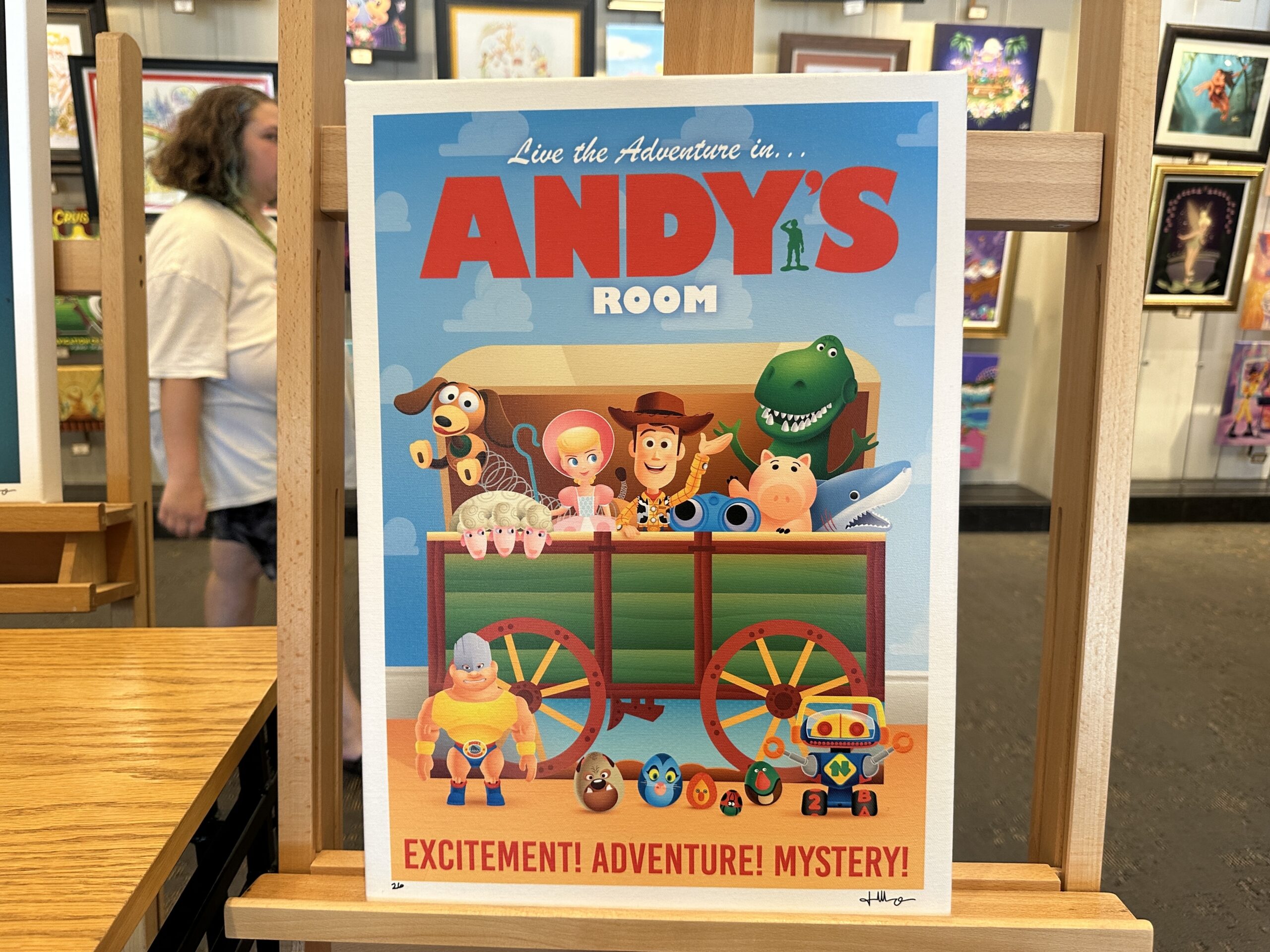 This piece in Jerrod Maruyama's style shows several of the original Toy Story characters in Andy's covered wagon toy box. Inside the open box are Slinky Dog, Bo Peep, Woody, Hamm, Rex, and more characters. At the top reads "Live the Adventure in...Andy's Room," and at the bottom says "Excitement! Adventure! Mystery!"