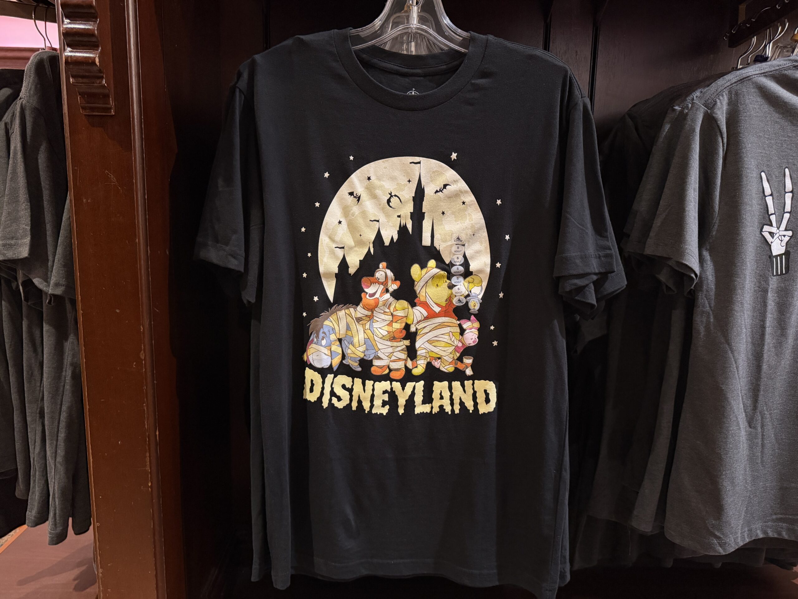 A black Fantasyland Castle outline is against a bright, full moon on this black T-shirt. Black bats fly around the castle and white stars surround the moon. In the foreground, Piglet, Pooh, Tigger, and Eeyore all dressed as mummies (they’re wrapped in toilet paper). “Disneyland” is in a spooky, wiggly ivory font at the bottom.