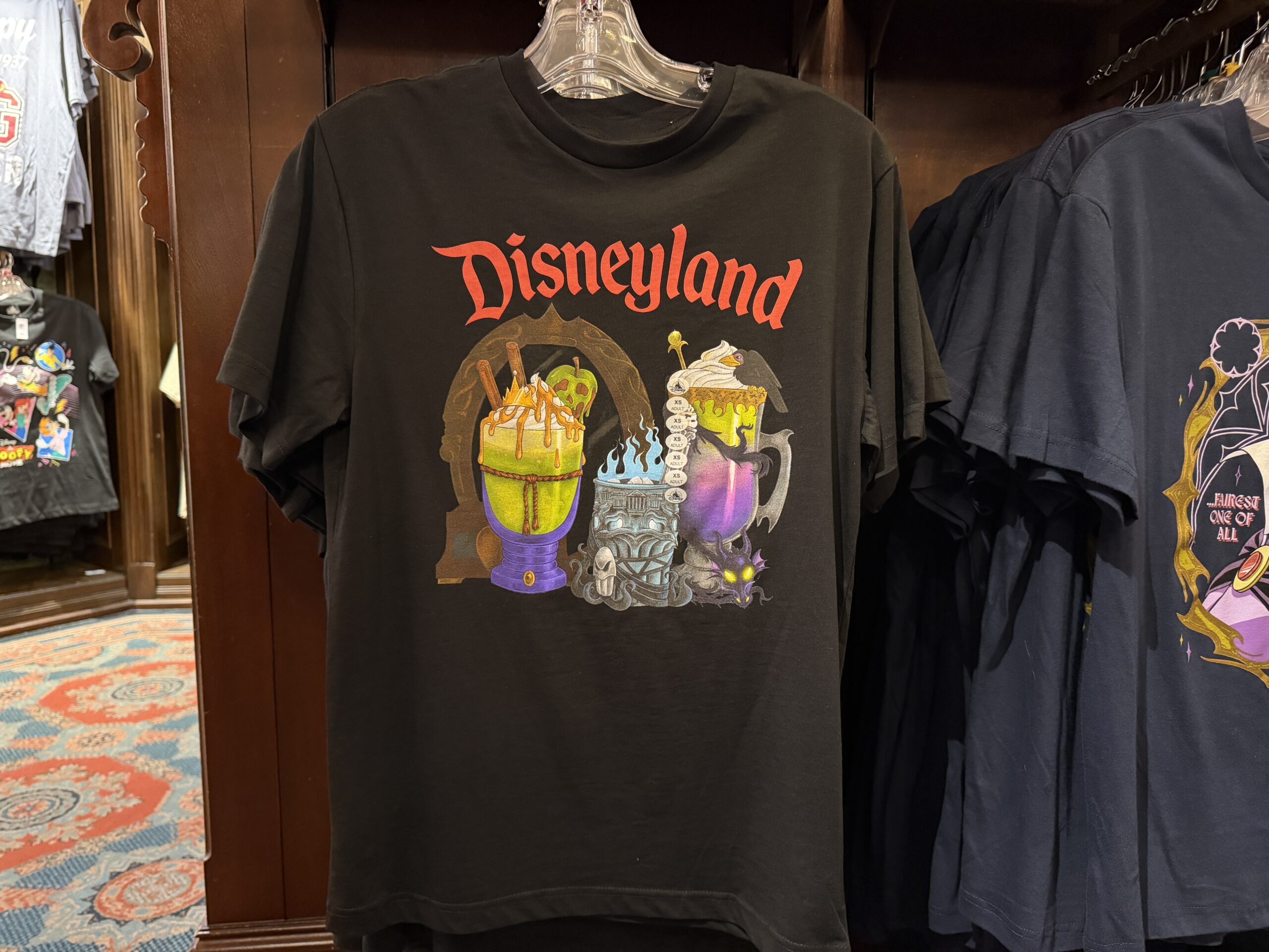 The Evil Queen, Hades, and Maleficent are all represented on this black T-shirt as different themed beverages. Each of the glasses is themed to match the inspirational villain. The Disneyland wordmark is in red at the top of the shirt.