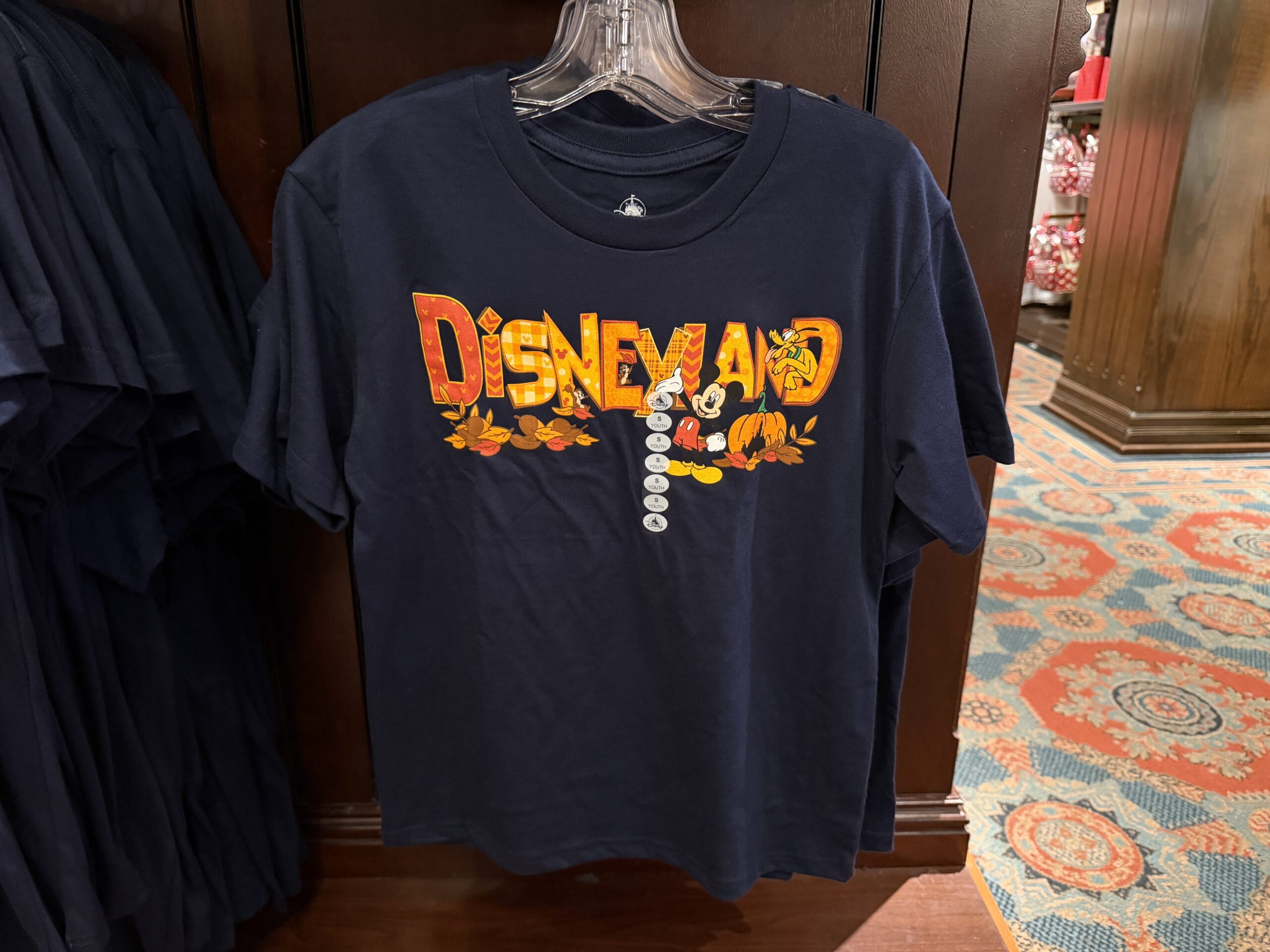 Mickey, Chip ‘n’ Dale, and Pluto are celebrating autumn on this autumnal shirt. The T-shirt is navy blue with “Disneyland” in patterned fall colors across the front. Acorns and autumn leaves are scattered beneath the wording, and a Fantasyland Castle Jack-o’-lantern is on the far right.