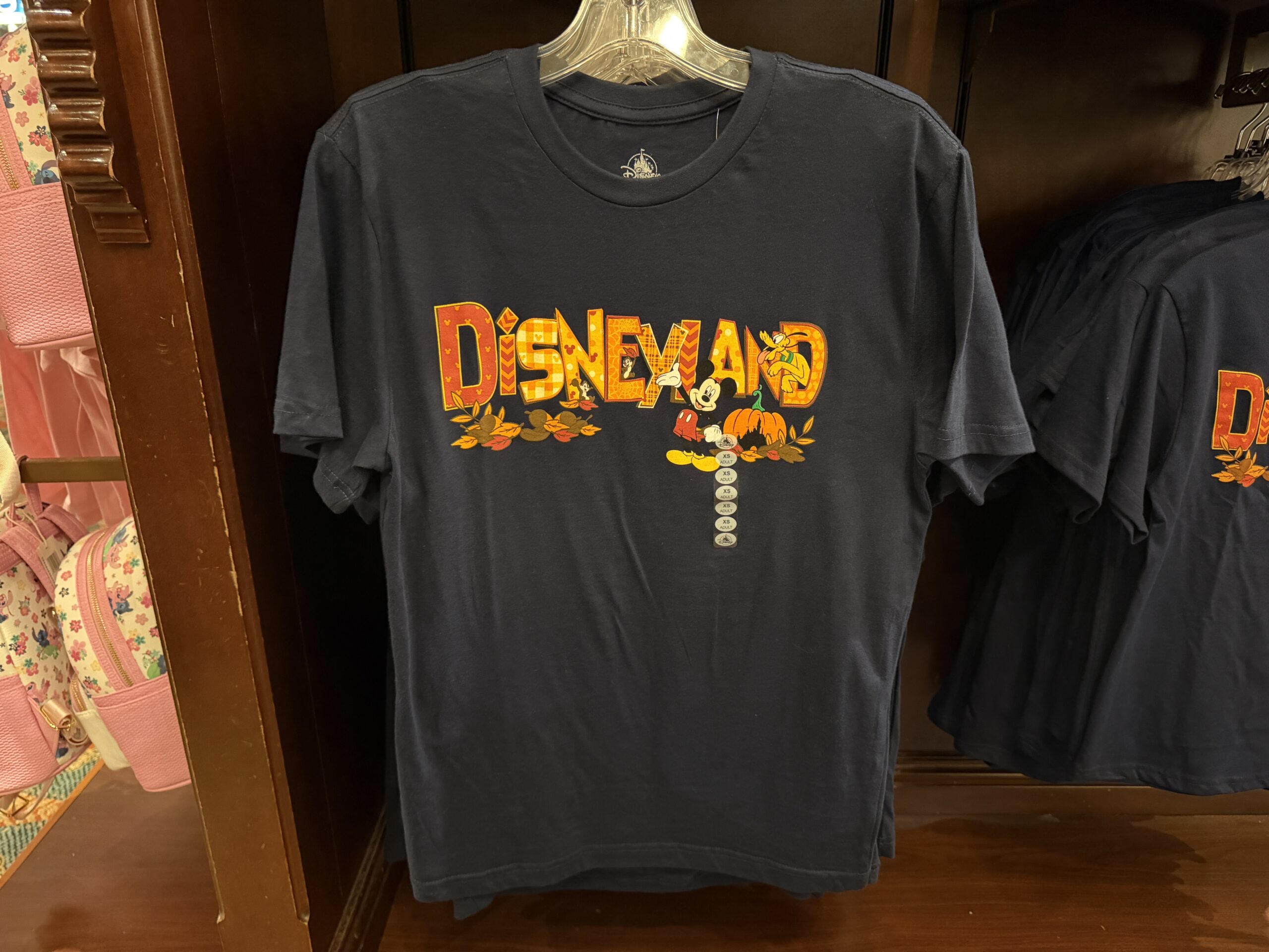 Mickey, Chip ‘n’ Dale, and Pluto are celebrating autumn on this autumnal shirt. The T-shirt is navy blue with “Disneyland” in patterned fall colors across the front. Acorns and autumn leaves are scattered beneath the wording, and a Fantasyland Castle Jack-o’-lantern is on the far right.