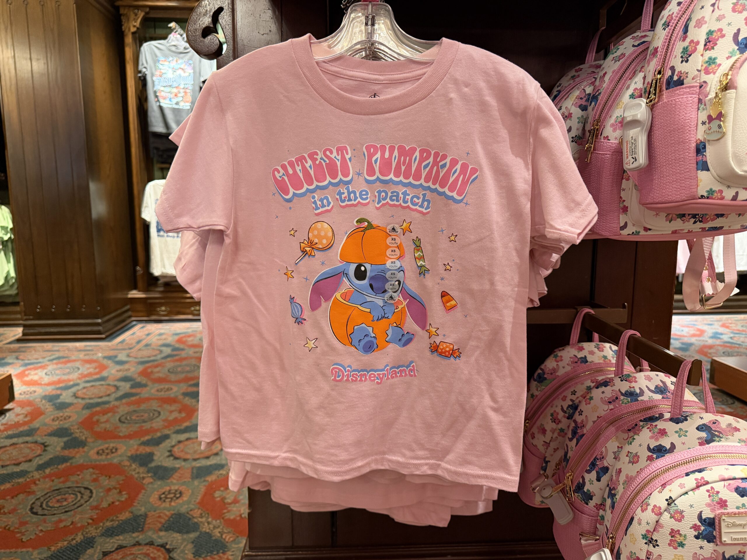 Stitch sits inside a pumpkin on this pink tee as Halloween candy surrounds him. "Cutest Pumpkin in the patch" is at the top in pink and blue text. "Disneyland" is at the bottom in the same style.