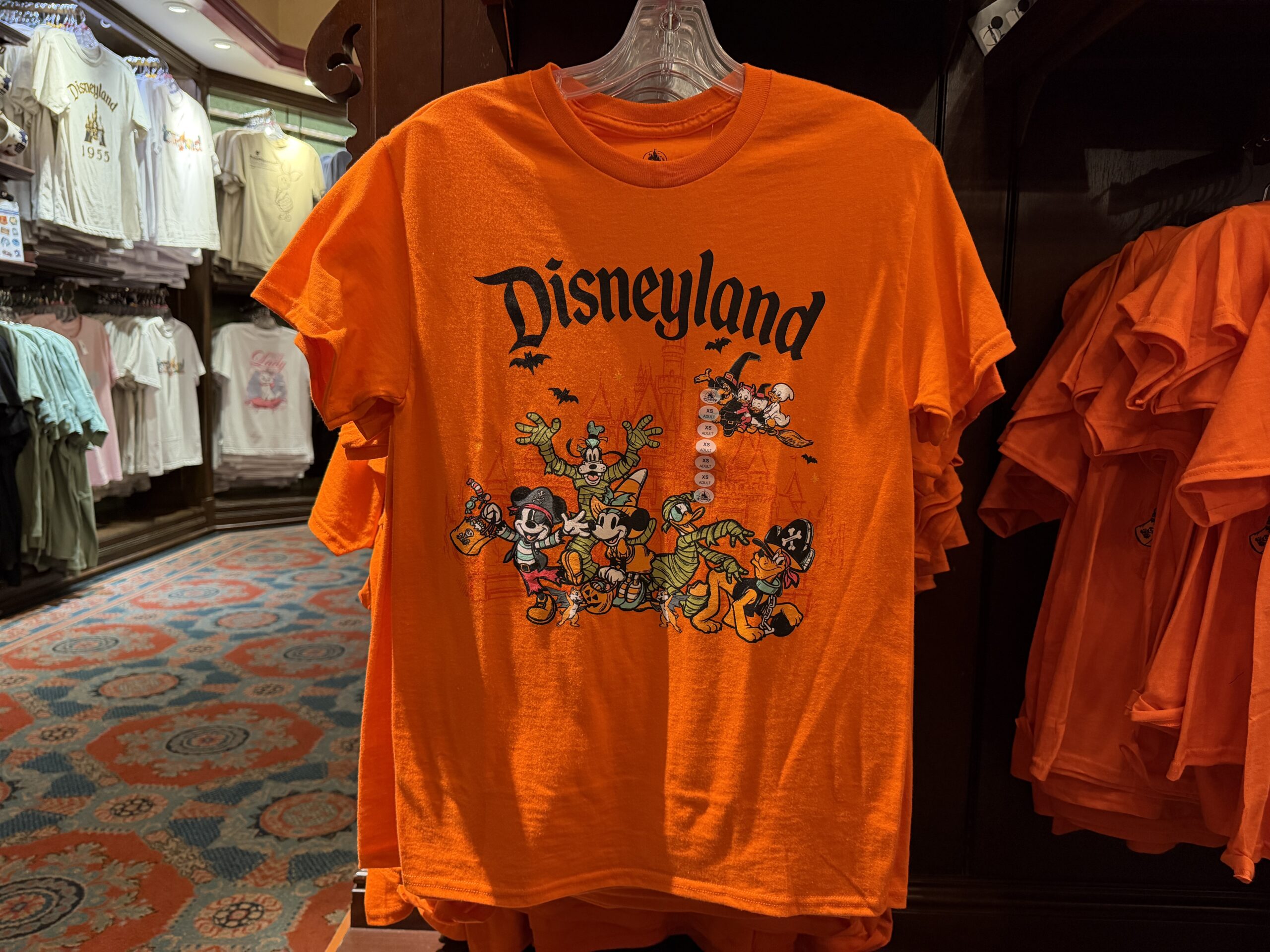 This orange tee has images of Mickey and his friends dressed in various costumes. Mickey is a pirate, Minnie is a witch, Goofy and Donald are both mummies, and Pluto is also a pirate. Behind them, bats fly and a witch carries Huey, Dewey, and Louie on a broom. The Disneyland wordmark is at the top in black.