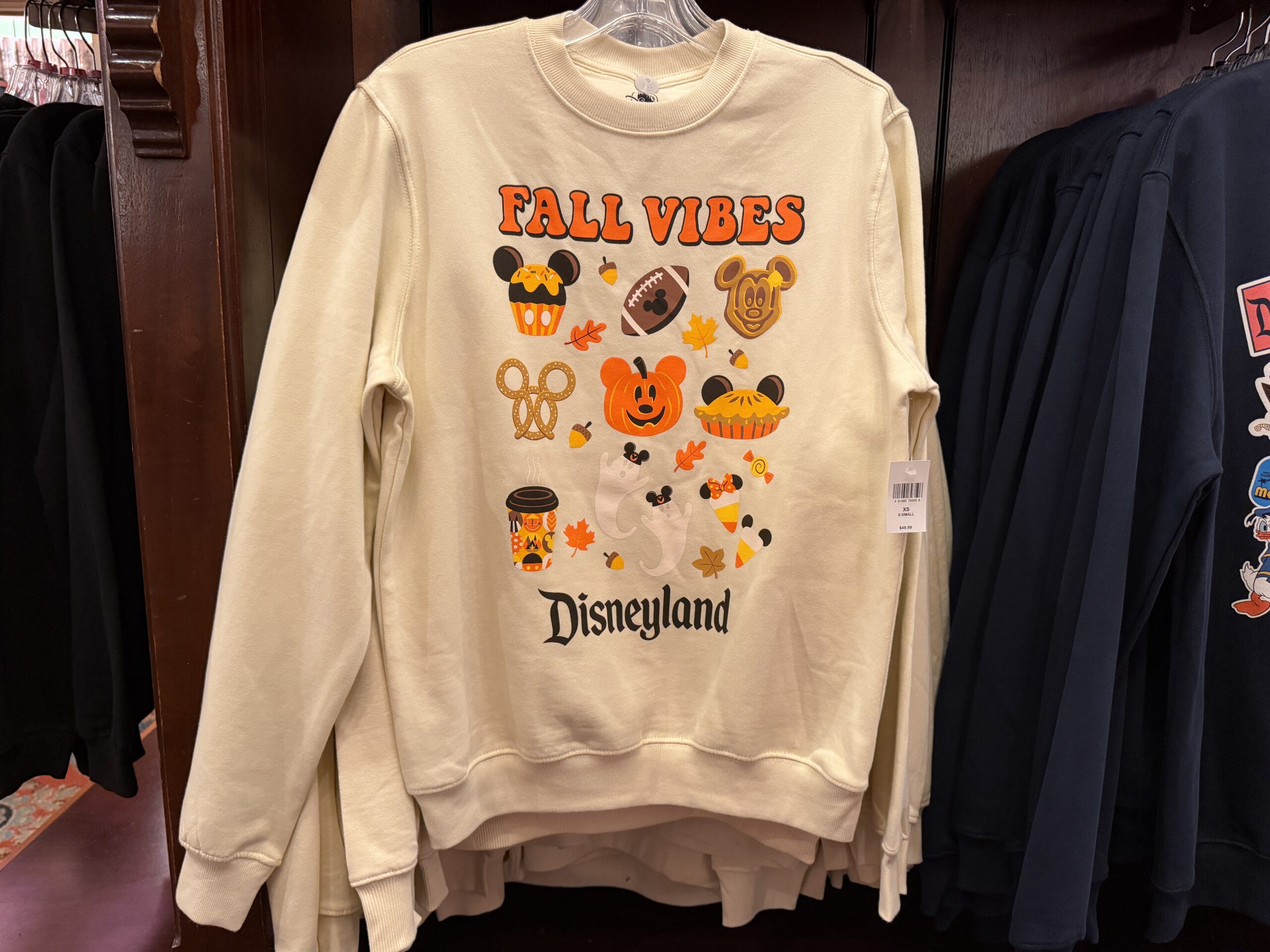 Celebrate fall with this beige tee featuring different autumnal icons. “Fall Vibes” is in orange lettering at the top, and “Disneyland” is in black at the bottom. The images (mostly shades of orange) include a Mickey cupcake, football, Mickey waffle, Mickey pretzel, Mickey Jack-o’-Lantern, Mickey pie, Disney coffee cup, ghosts wearing Mickey ears, candy corn-inspired ice cream cones, leaves, and acorns.