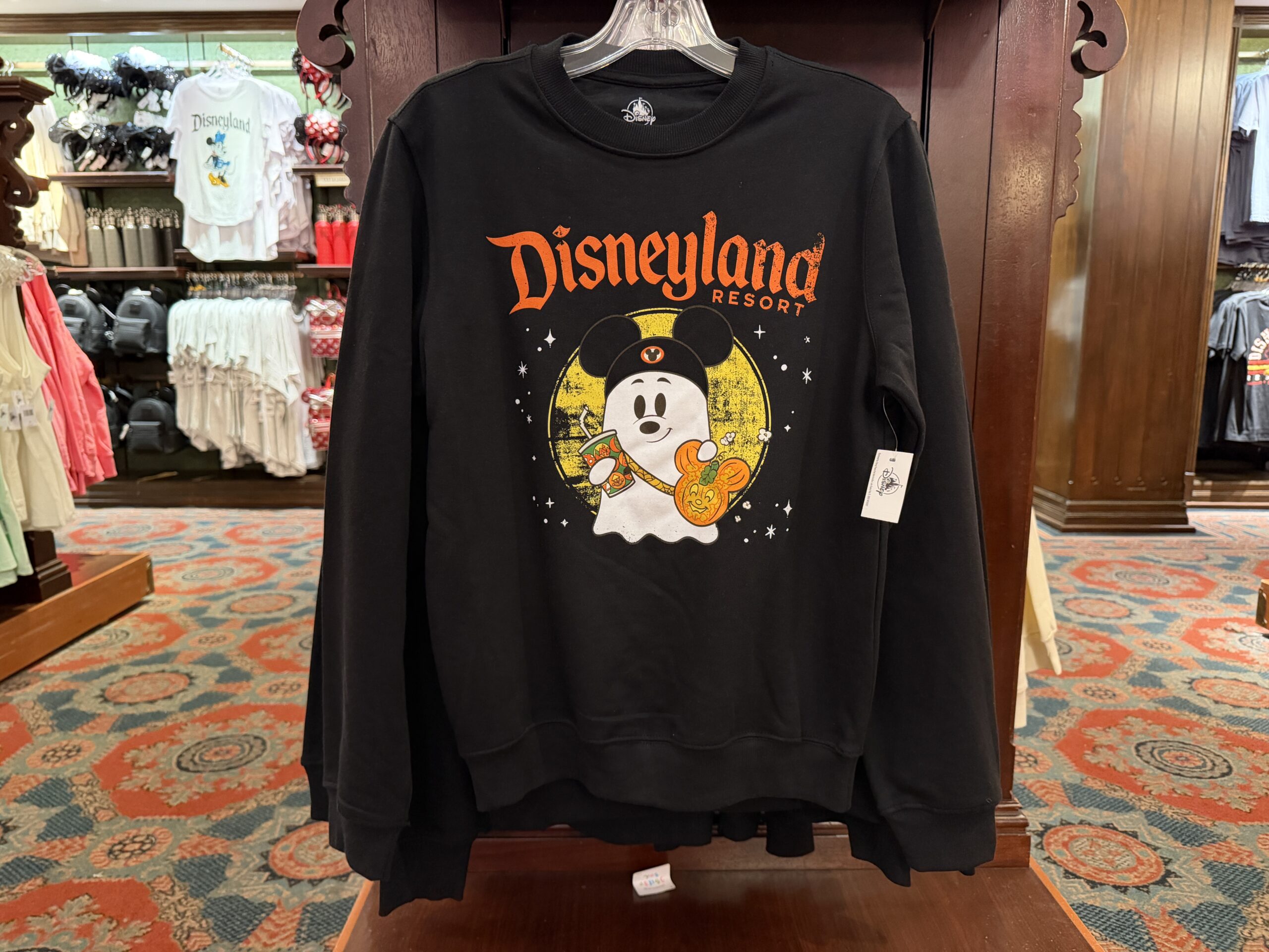 A black sweatshirt features a ghost wearing a Mickey ear hat. The ghost is set against a yellow circle with faux weathering. White stars are around the image. “Disneyland Resort” is in orange lettering above the ghost. It’s carrying a Disney soda cup and a Jack-o’-Lantern popcorn bucket.