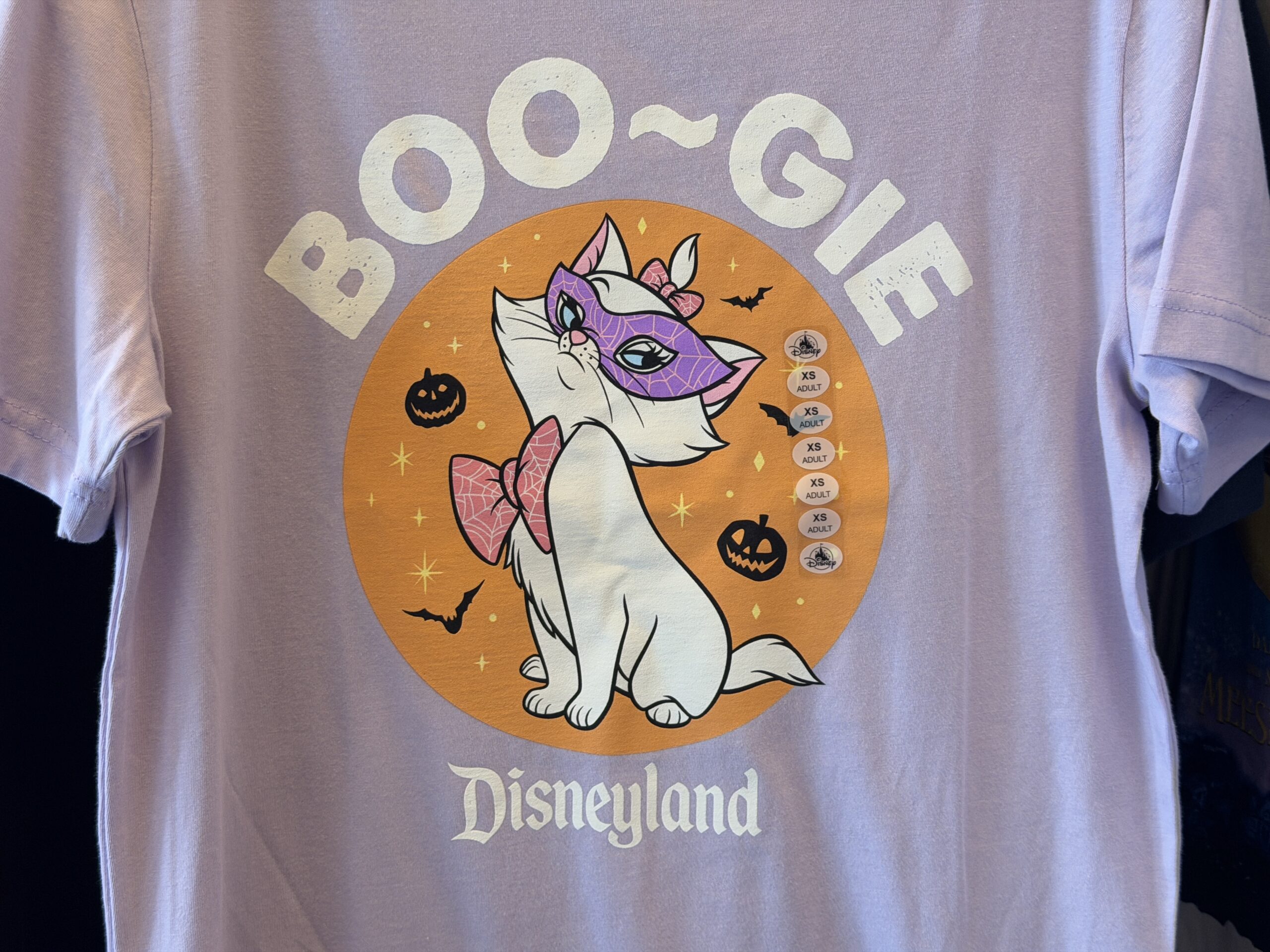Marie is in the spotlight on this lavender shirt. She is against an orange, circular background patterned with stars, Jack-o’-lanterns, and bats. Marie is wearing a purple mask with a white cobweb pattern on it, which matches the pattern on her pink hair and neck bows. Above her head is the word “Boo-gie,” likely a pun on “bougie,” in white. The Disneyland wordmark is also in white at the bottom.