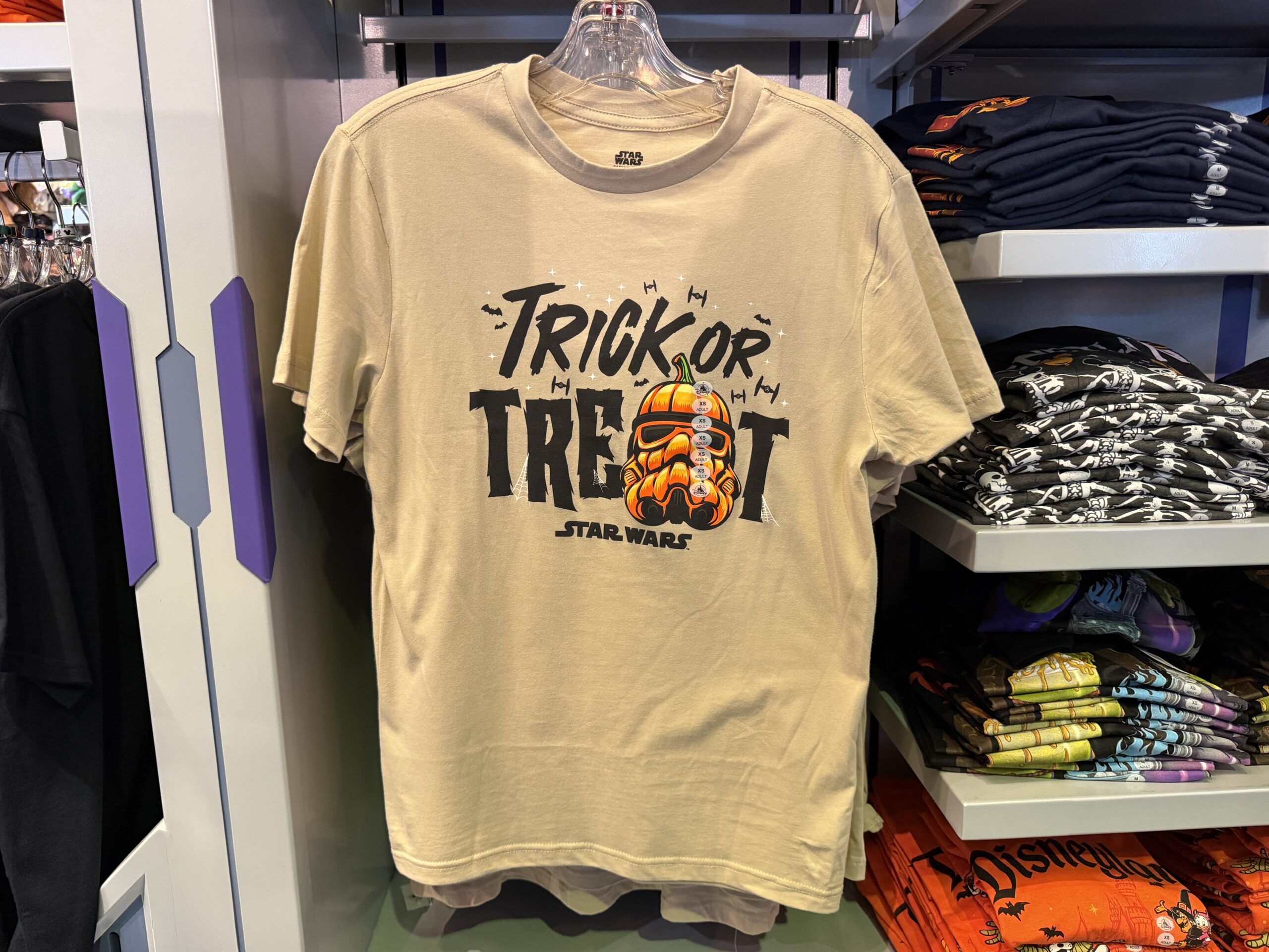 A Stormtrooper helmet has been turned into a shaped pumpkin on the front of this light gray T-shirt. It says “Trick or Treat” on the front, though the “A” has been replaced by the Stormtrooper helmet. The Star Wars wordmark is at the bottom in black while black bats and TIE Fighters soar above amongst white twinkly stars.