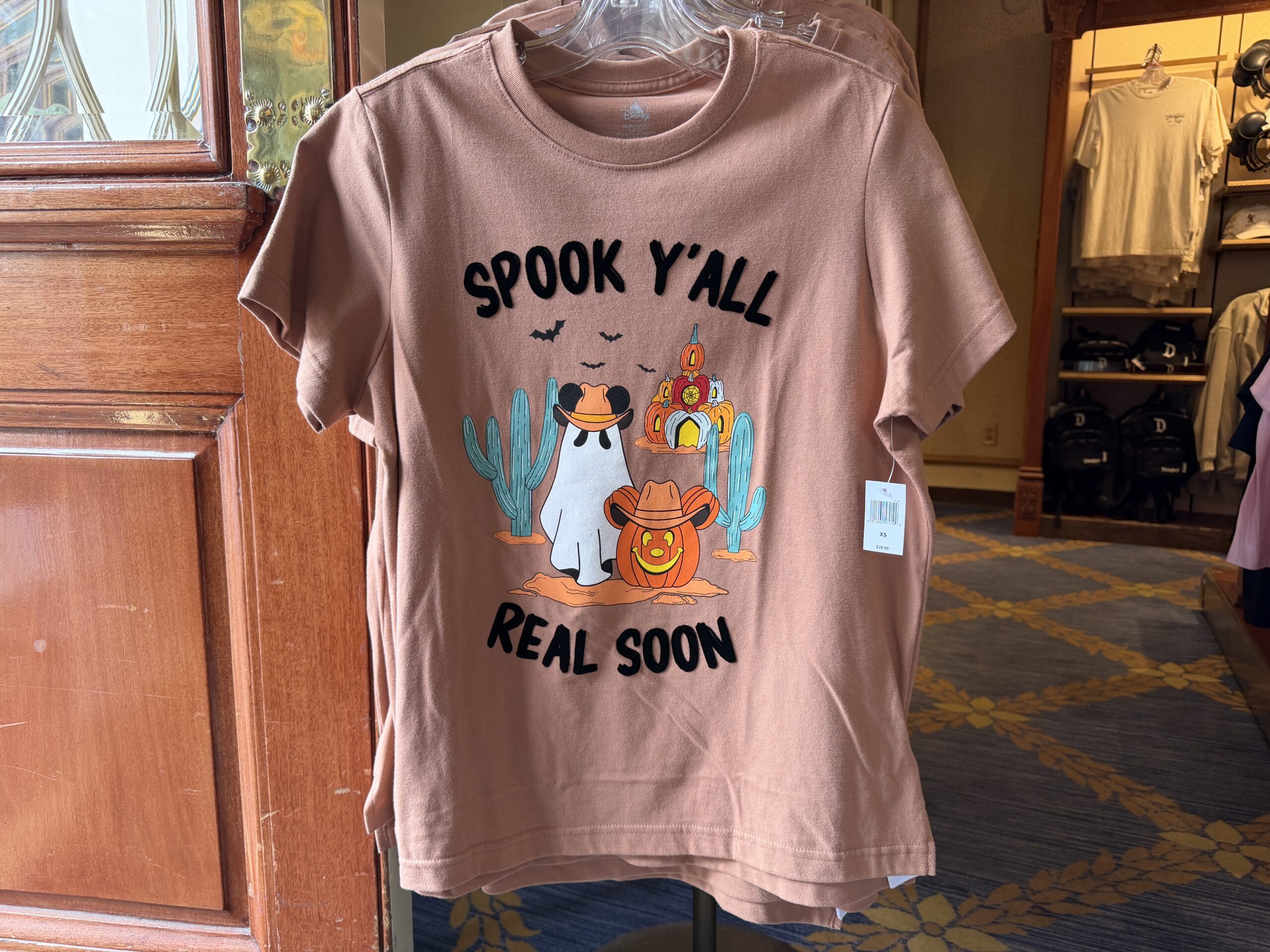 This light orange tee has "Spook Y'all Real Soon" in black text. It surrounds an image of a Mickey ghost, Mickey Jack-o’-Lantern, pumpkin castle, and two cacti. Bats fly around in the background.