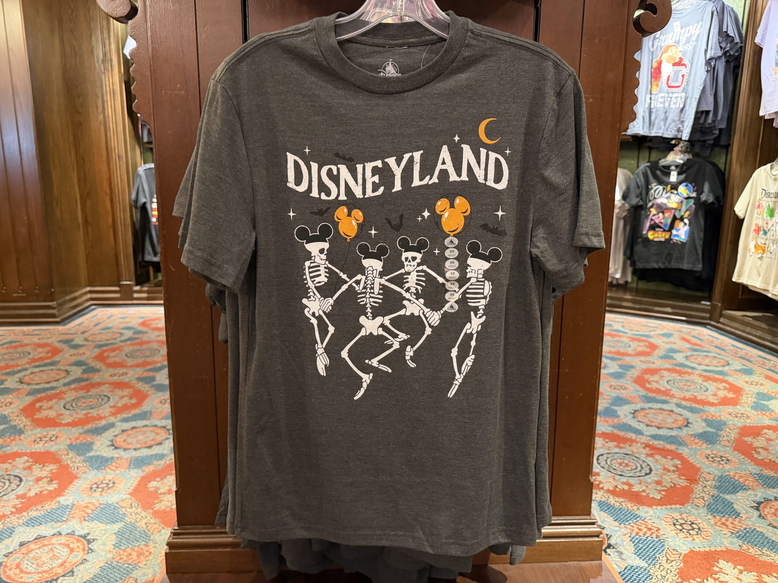 The four skeletons from the 1929 short “The Skeleton Dance” are all wearing Mickey ear hats, and two are holding orange Mickey ear balloons on this dark gray T-Shirt. White star sparkles, black bats, an orange moon, and “Disneyland” in white are above them.