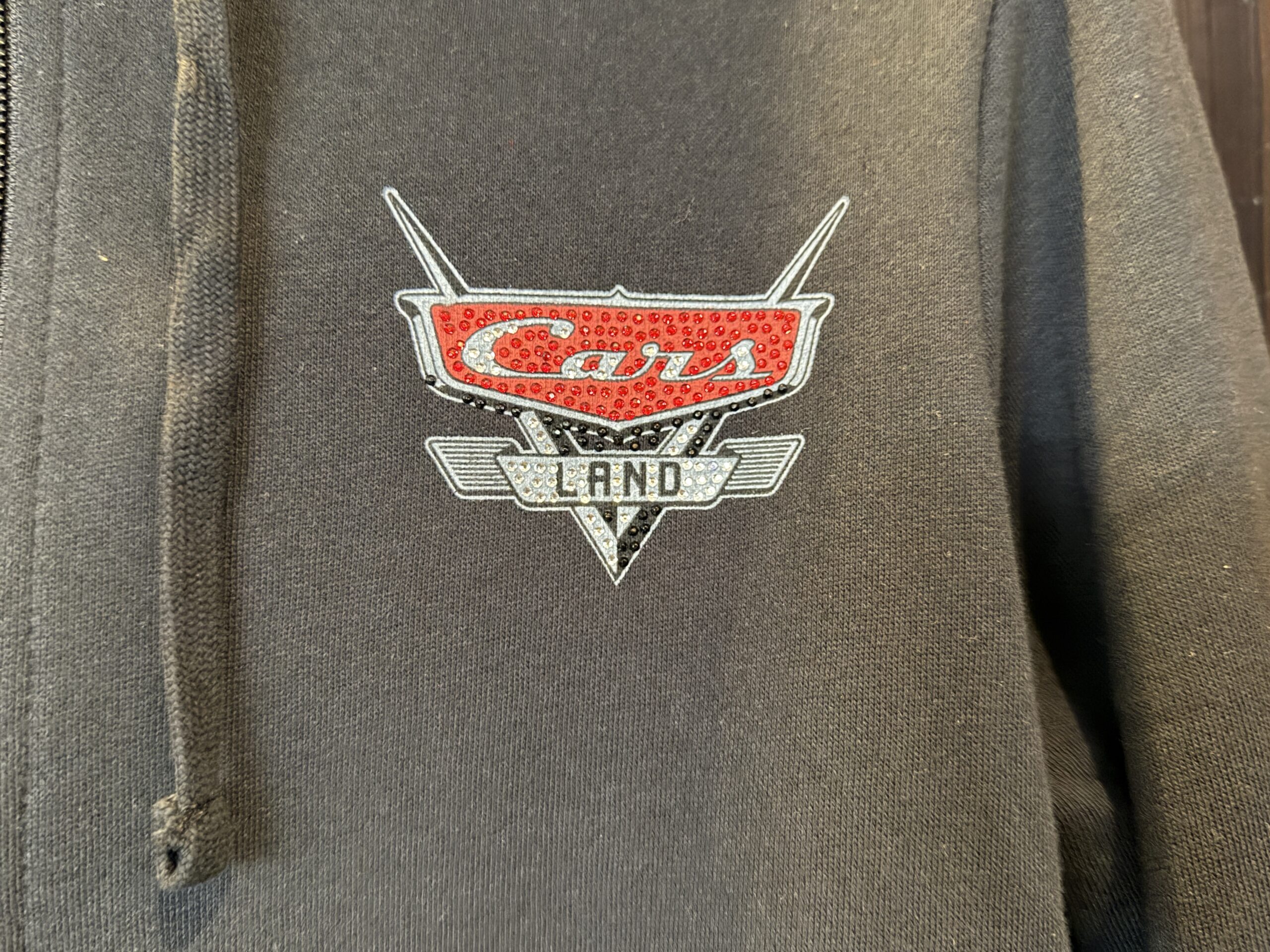 The black zip-up hoodie has a front pocket. On the chest is the Cars Land logo in black, white, and red. Most of the logo is bedazzled.