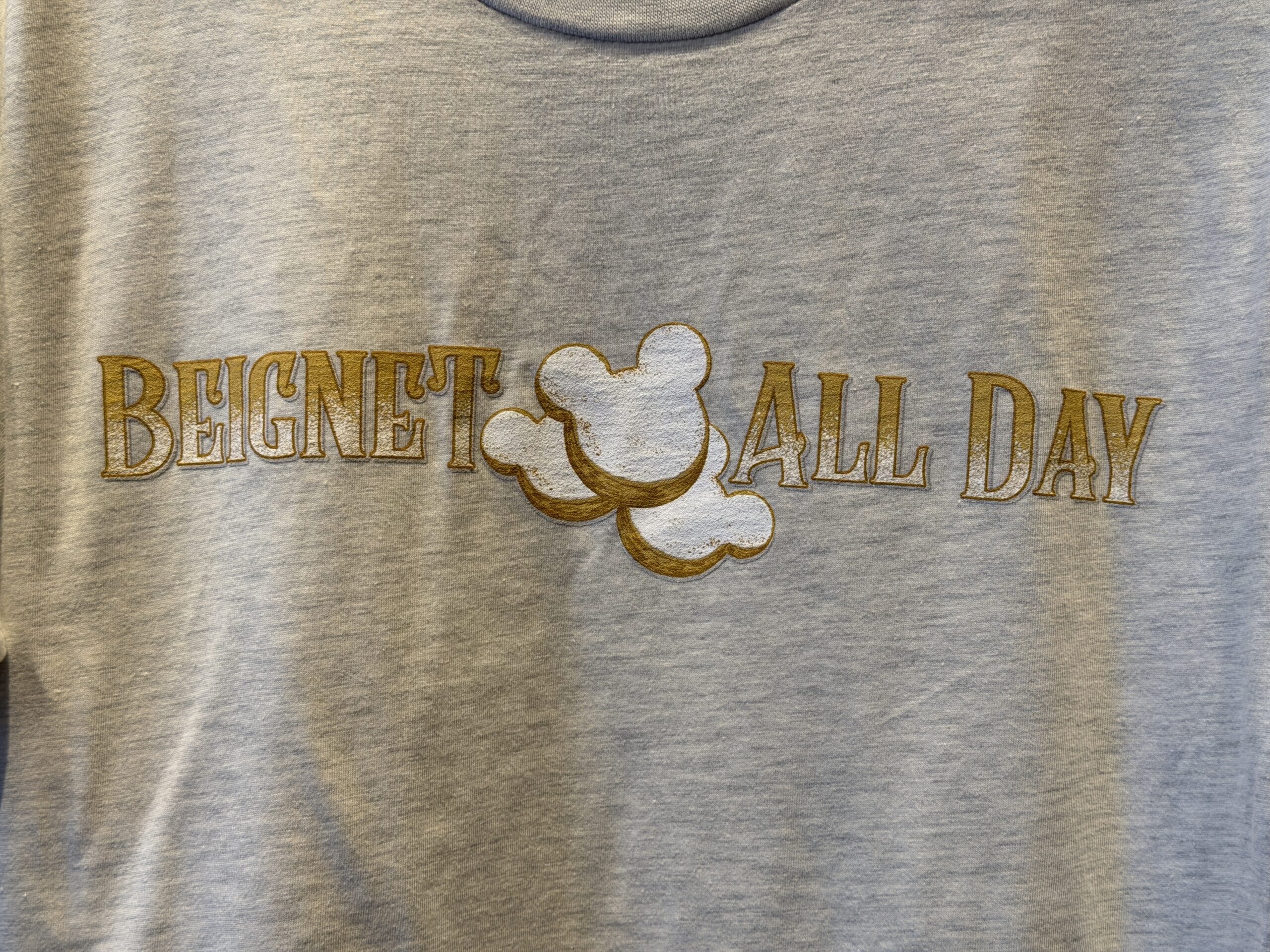 The gray T-shirt has "beignet all day" text in ombre dusted beige and white text. Three beignets are placed in the center with powdered dusting.