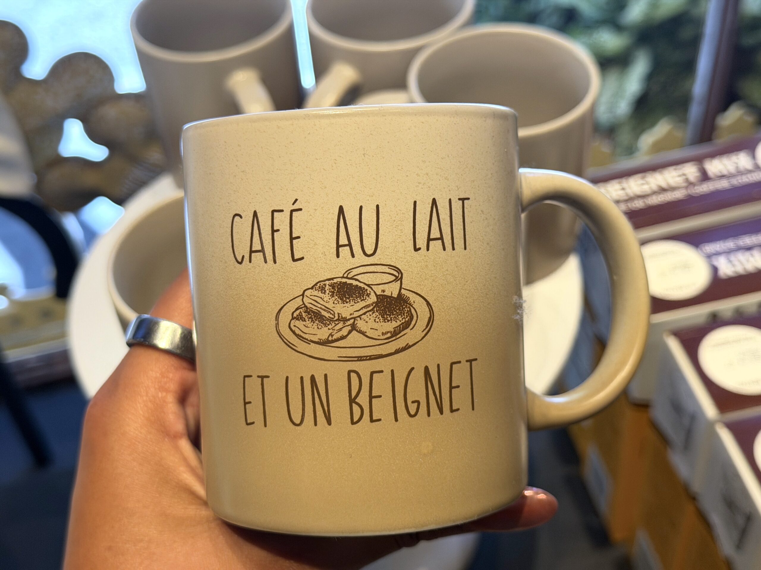 This ombre white and beige-colored mug has "Café au Lait" text at the top, a plate of beignets in the center, and "Et un beignet" at the bottom.