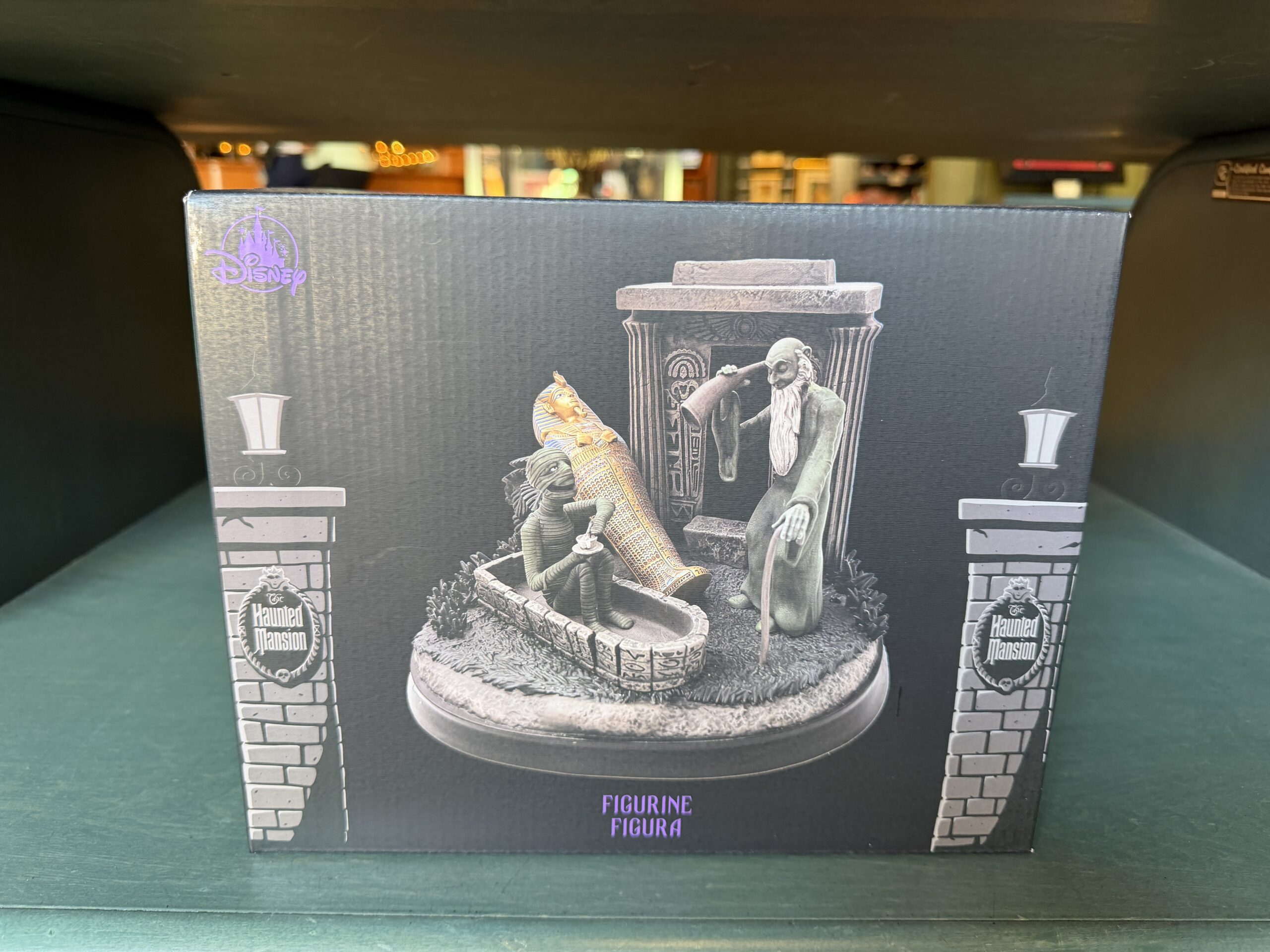 According to the back of the box, the musical figurine plays the song "Grim Grinning Ghosts." The back of the box has green and black vertical striped wallpaper and The Haunted Mansion logo in the center. The front shows the musical figurine between two entry columns for the mansion.