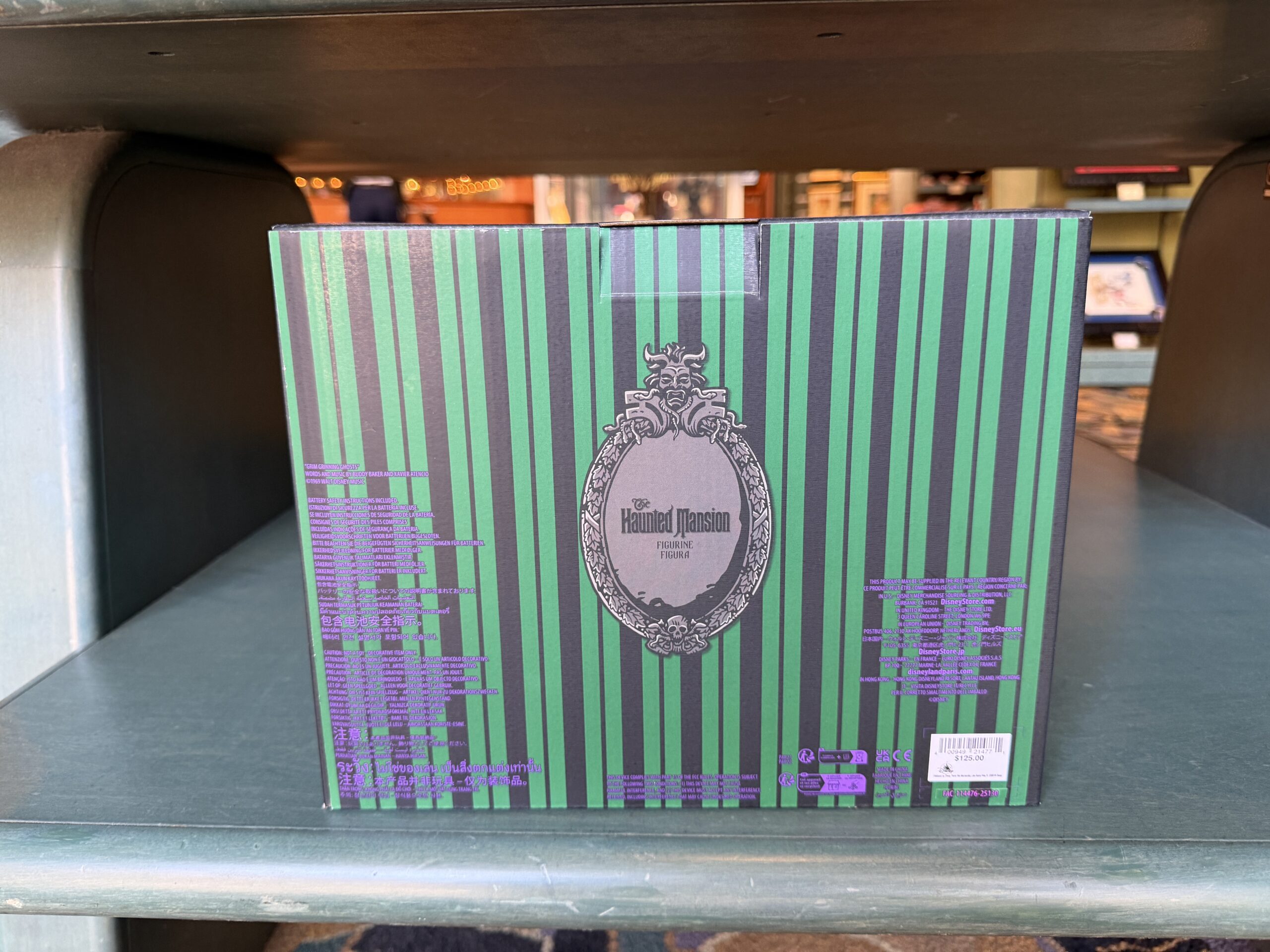 According to the back of the box, the musical figurine plays the song "Grim Grinning Ghosts." The back of the box has green and black vertical striped wallpaper and The Haunted Mansion logo in the center. The front shows the musical figurine between two entry columns for the mansion.