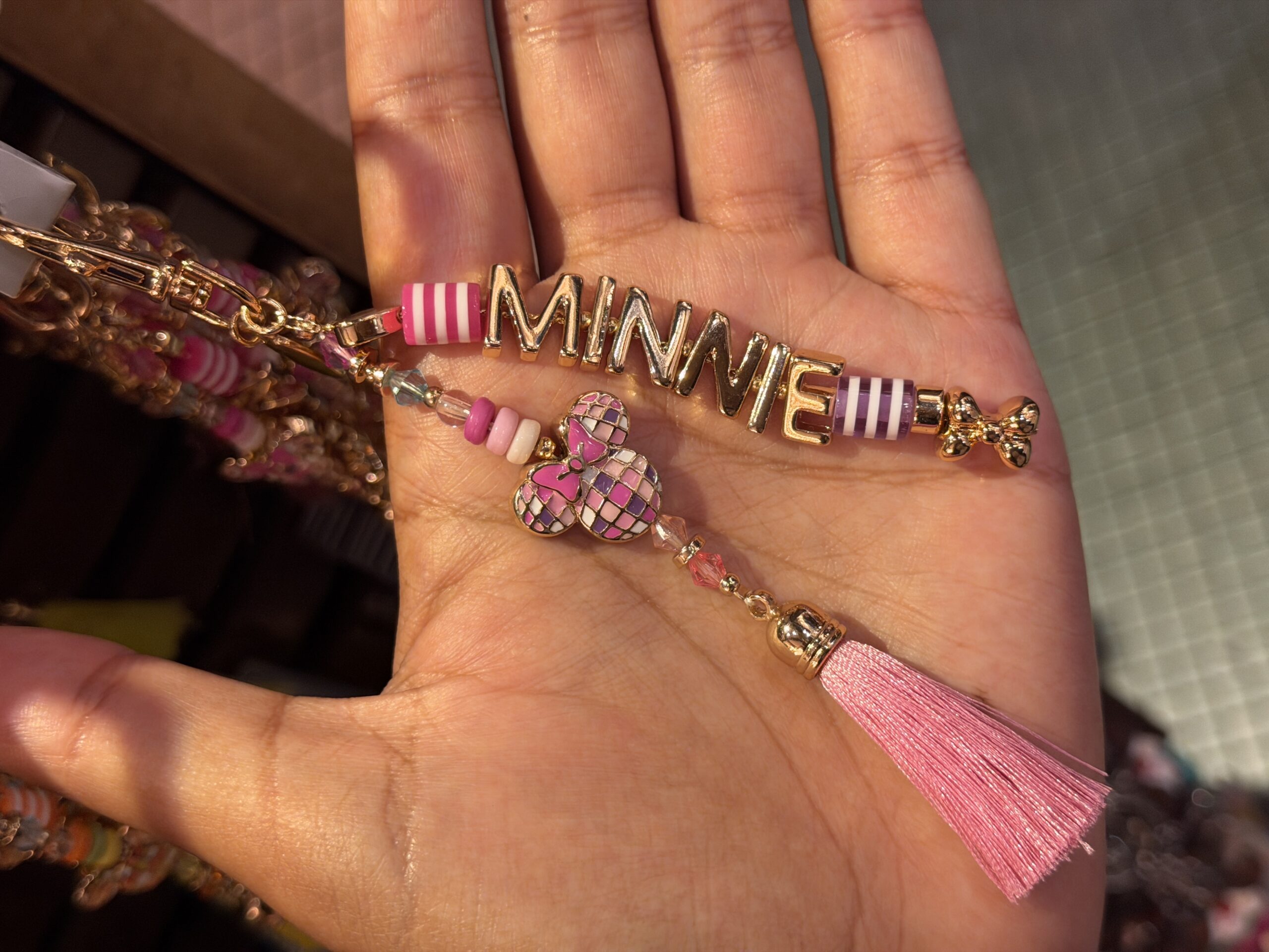 This charm has two strands of pink and gold beads. One spells out Minnie's name. On the other is a disco ball-like charm shaped like Minnie's head with a bow. A pink tassel dangles at the bottom.