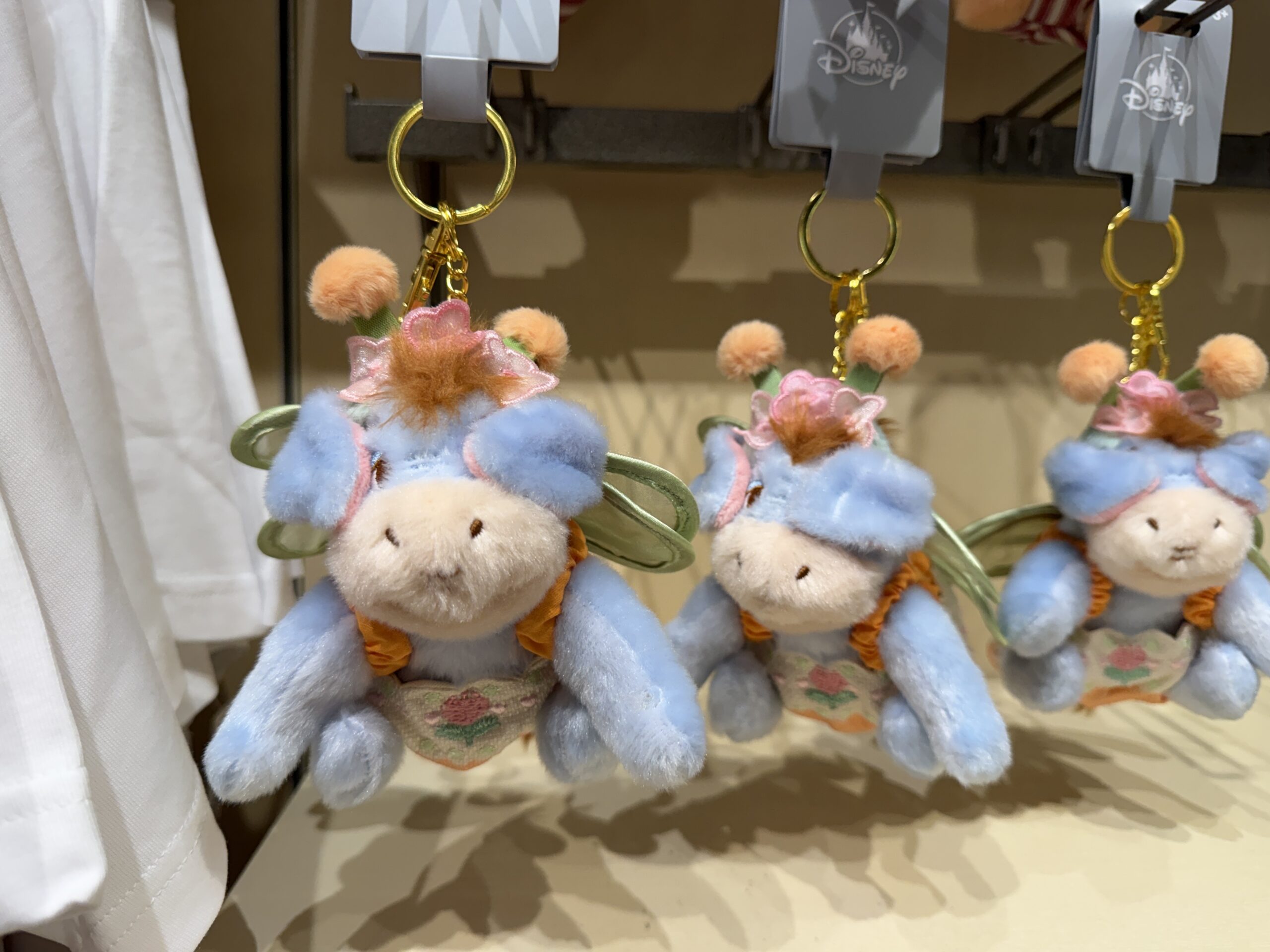 This plush depicts Eeyore dressed as a bee with fluffy orange antennae and green wings attached to a floral jumpsuit. He dangles from a goldtone chain on a split ring with a lobster claw clip.