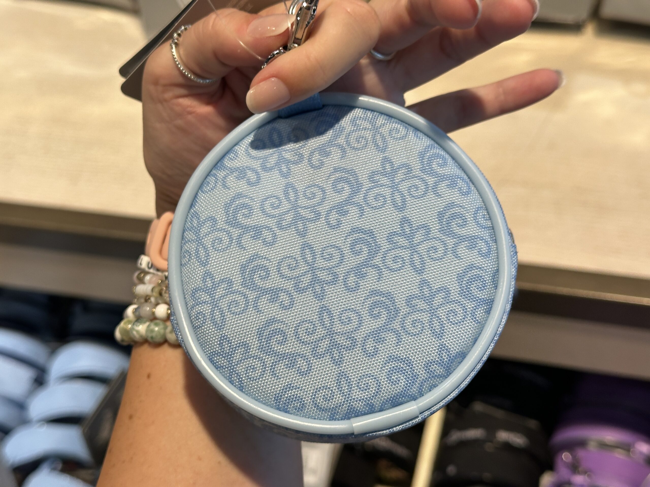 The front of this circular coin purse says "Living in a Fantasyland" with a blue Fantasyland Castle behind it. Silver stars are strewn throughout, and the Walt Disney World wordmark is on the bottom. The back is light blue and patterned with a darker blue filigree pattern that matches the castle on the front.