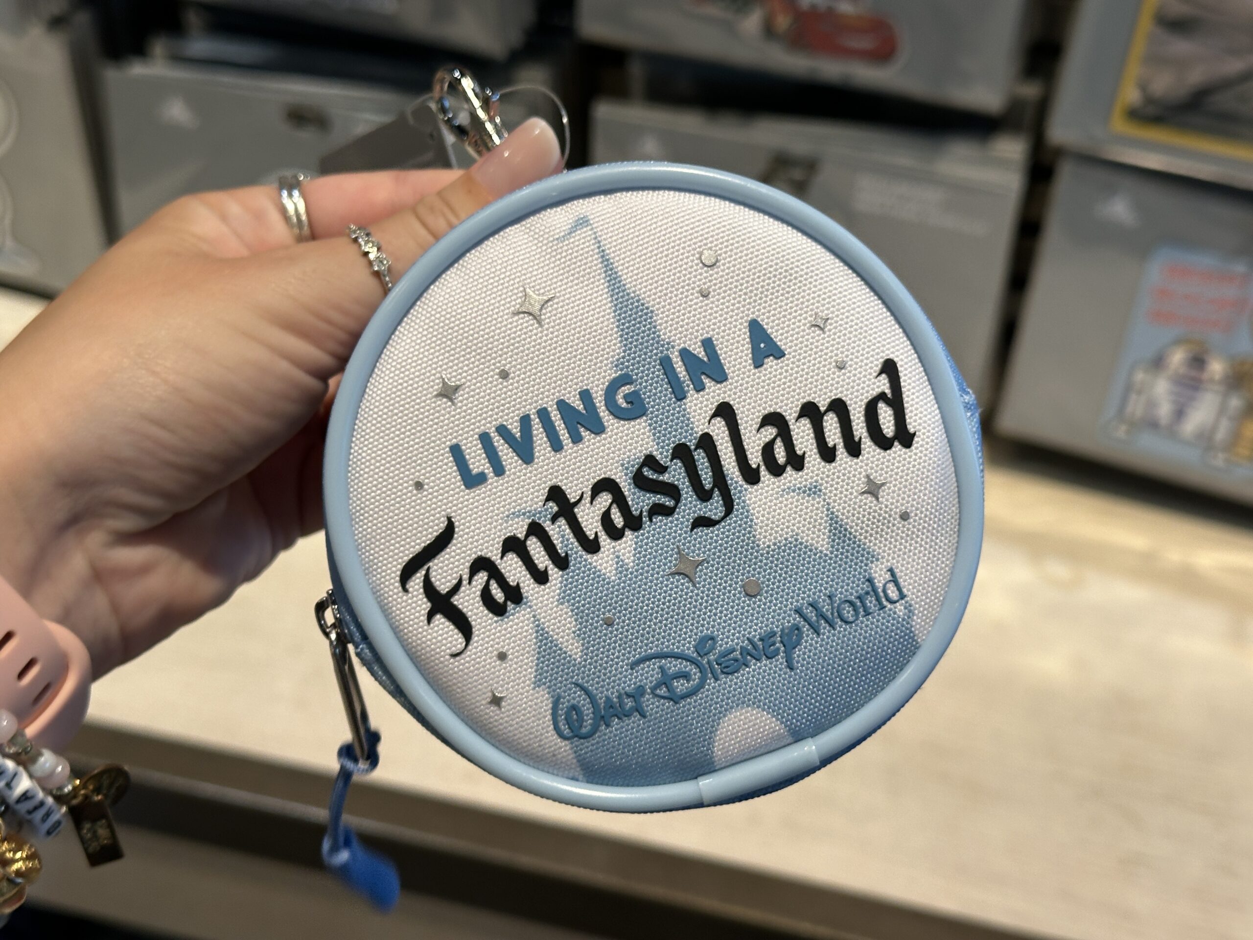 The front of this circular coin purse says "Living in a Fantasyland" with a blue Fantasyland Castle behind it. Silver stars are strewn throughout, and the Walt Disney World wordmark is on the bottom. The back is light blue and patterned with a darker blue filigree pattern that matches the castle on the front.