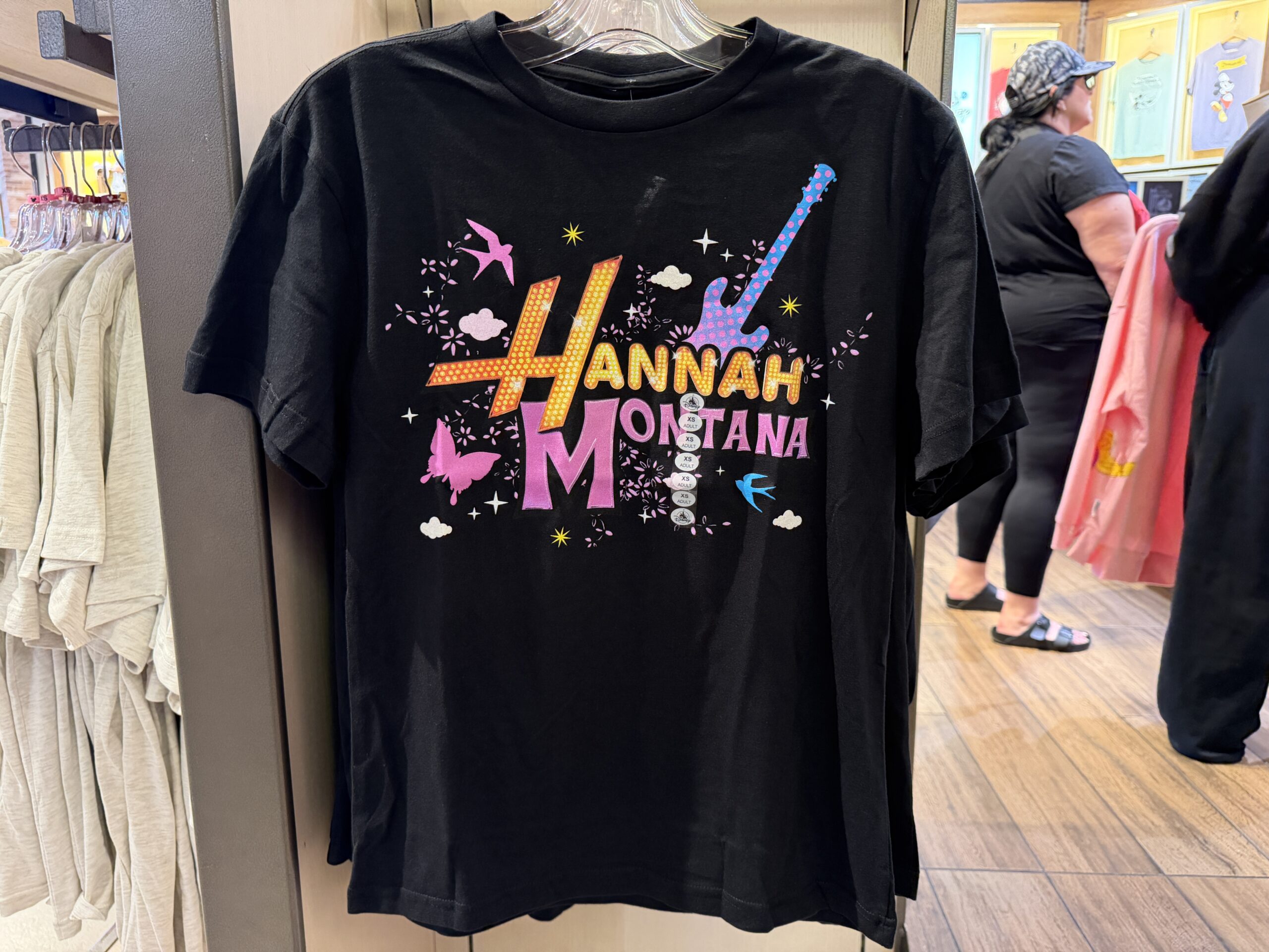 The Hannah Montana logo is in yellow-orange and pink against a black tee. Iconography includes butterflies, birds, and a guitar.