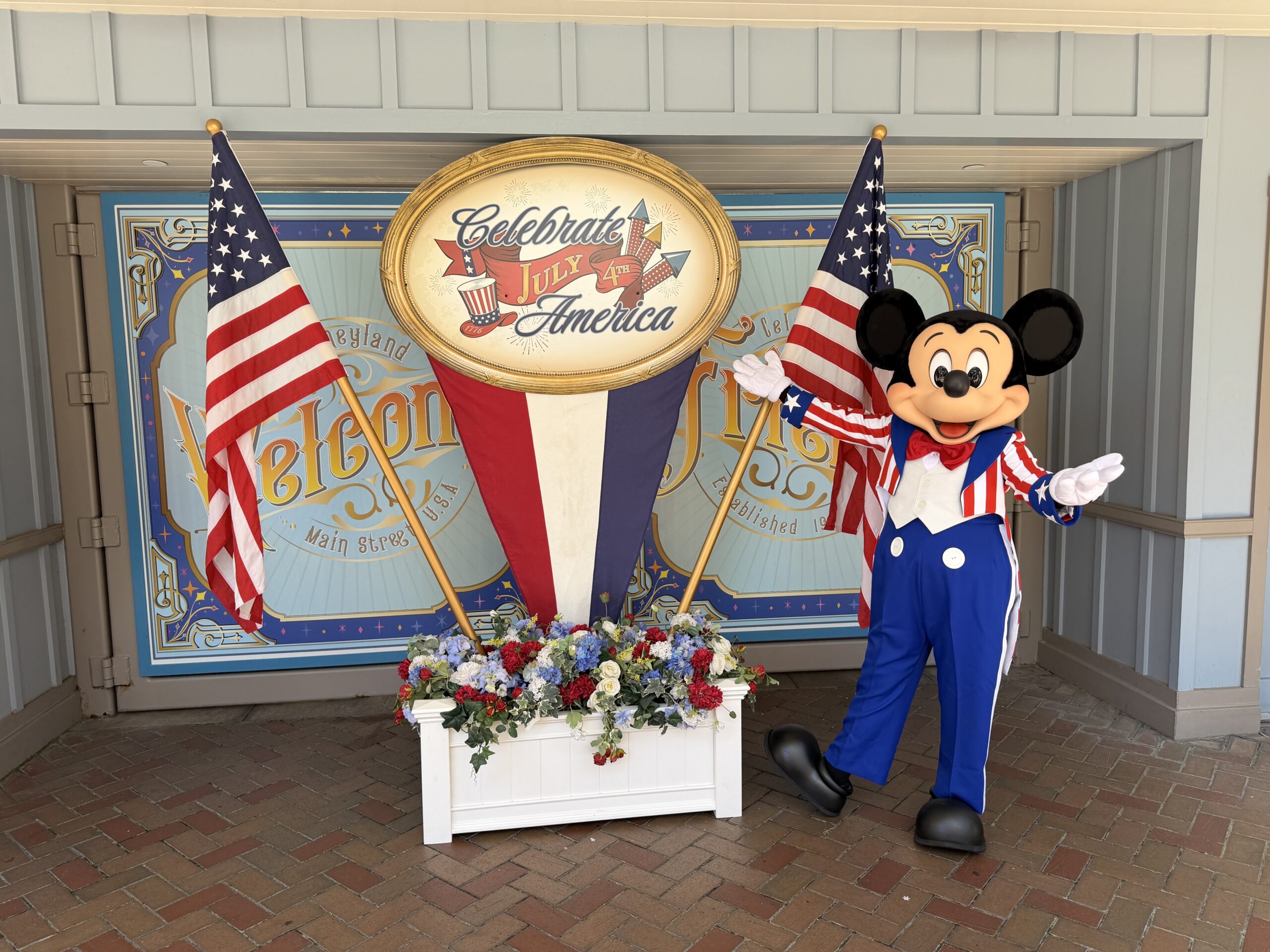 Mickey is wearing a red and white vertical-striped suit jacket with a blue collar and cuffs with stars around his wrists. He's got a red bowtie and blue pants. Beside him is a "Celebrate America" and "4th of July" standee. Red, white, and blue flowers with foliage are in a white flower box below. The standee has an American flag on either side.