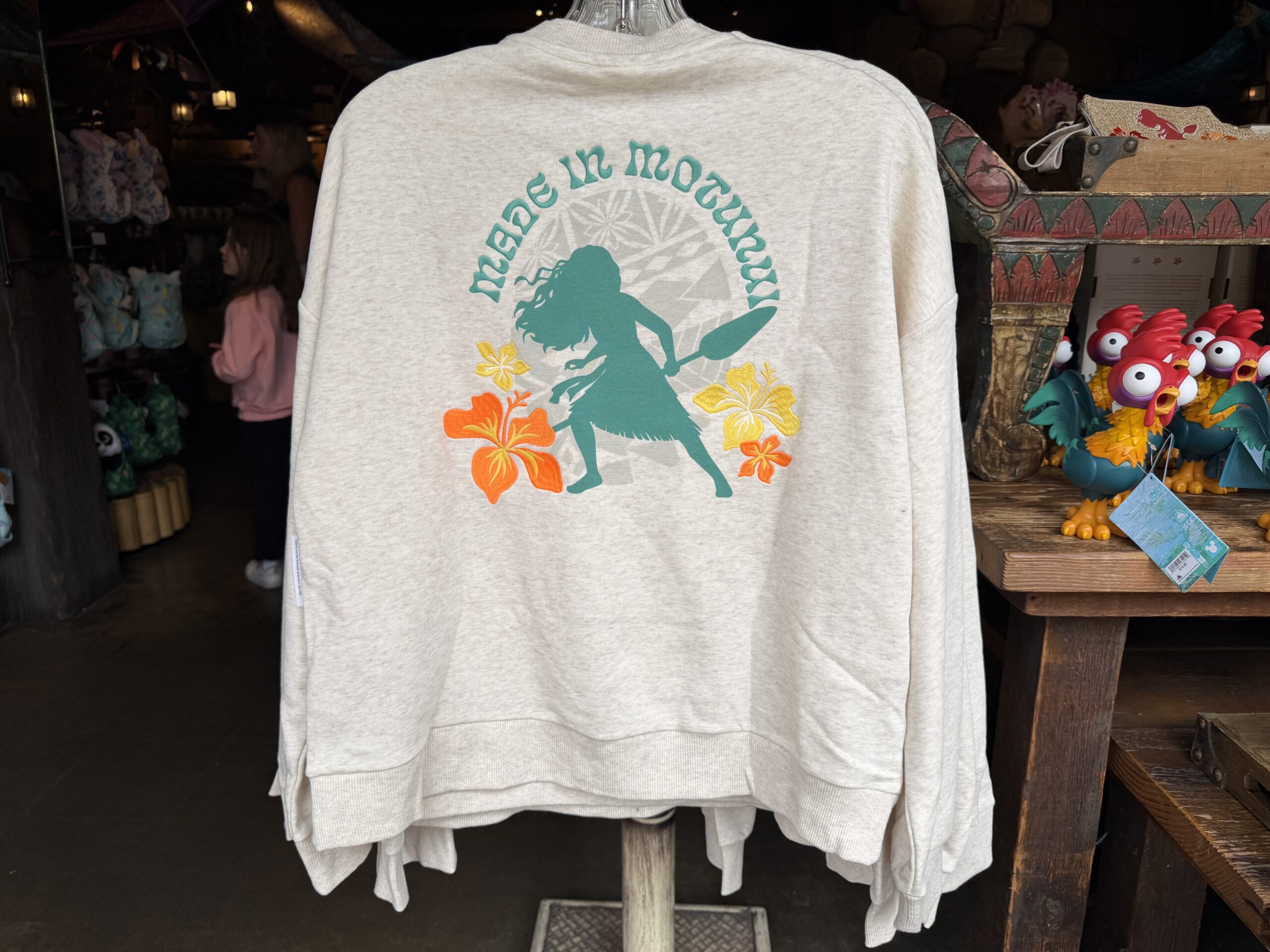 On the back, we see Moana without her boat, but holding her oar. The words "Made in Motunui" are above her, and she has a yellow and orange tropical flower on either side of her.