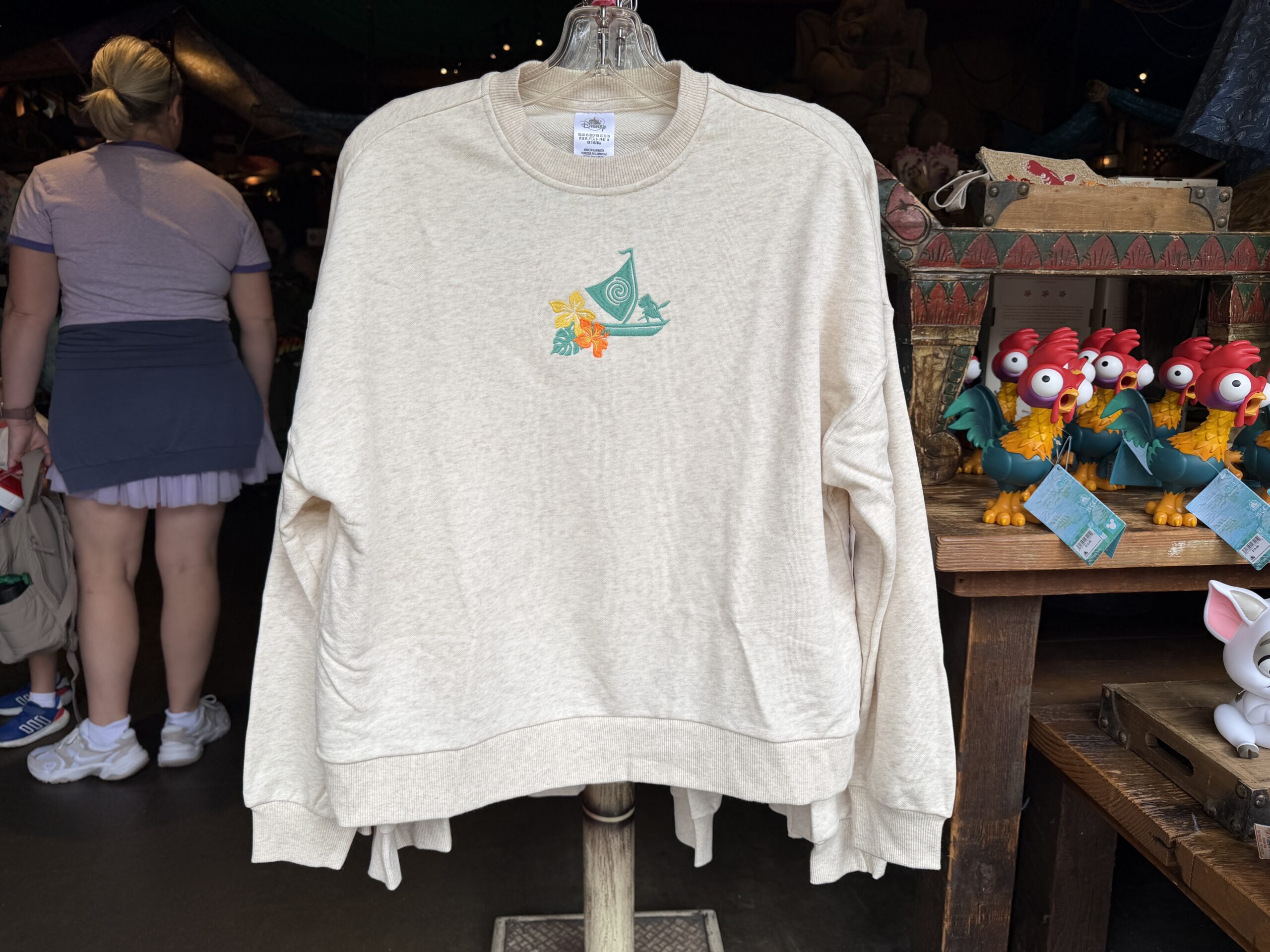 This cream-colored pullover sweatshirt has an embroidered aqua Moana aboard her boat sailing the sea. A pair of tropical flowers is beside it.