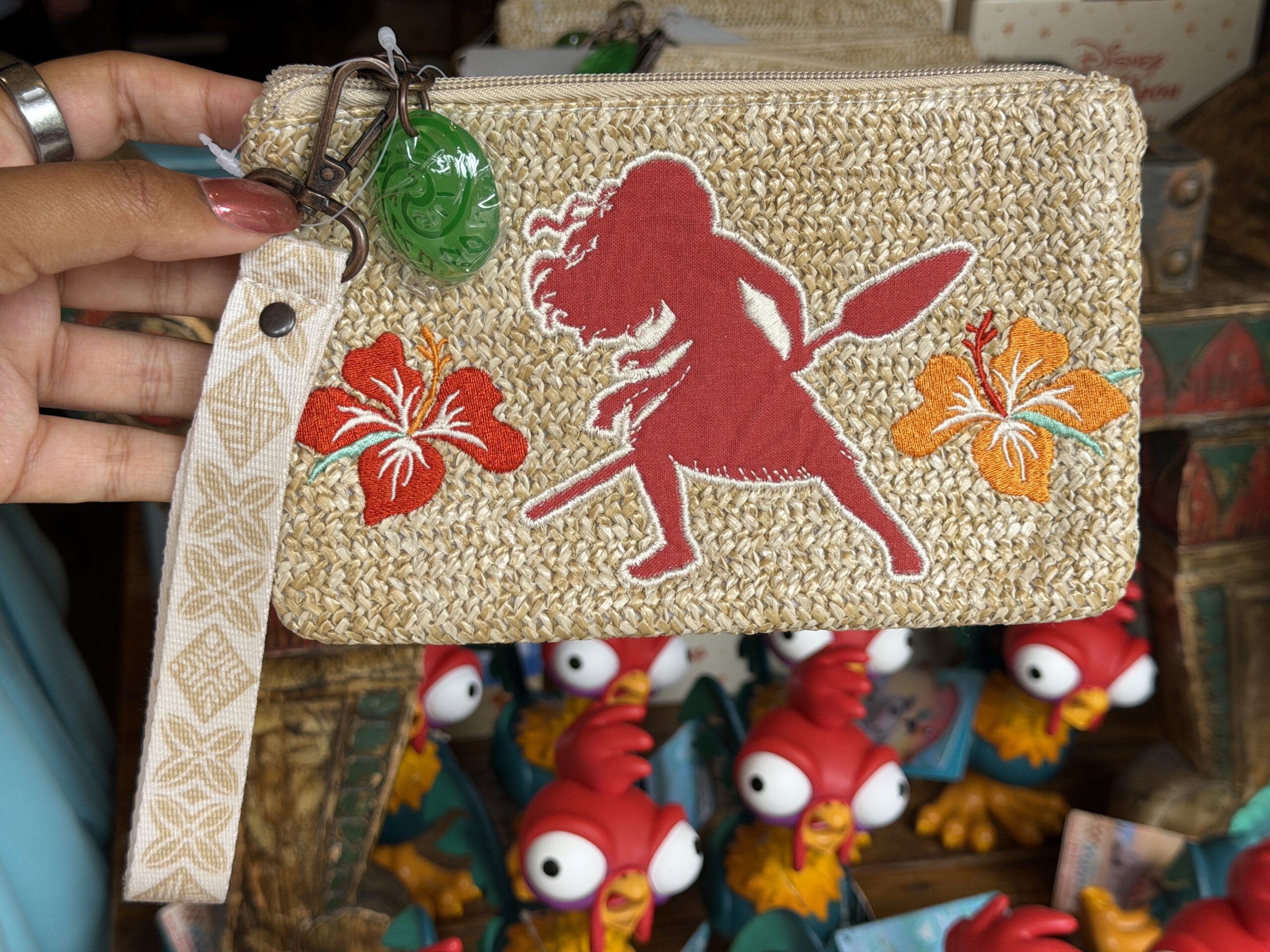 This rattan wristlet has a patch of Moana holding her oar in a burnt orange color. A matching flower is on her right side, and a yellow flower is on her left. A charm of The Heart of TeFiti is on the lobster claw clip of the zippered bag.