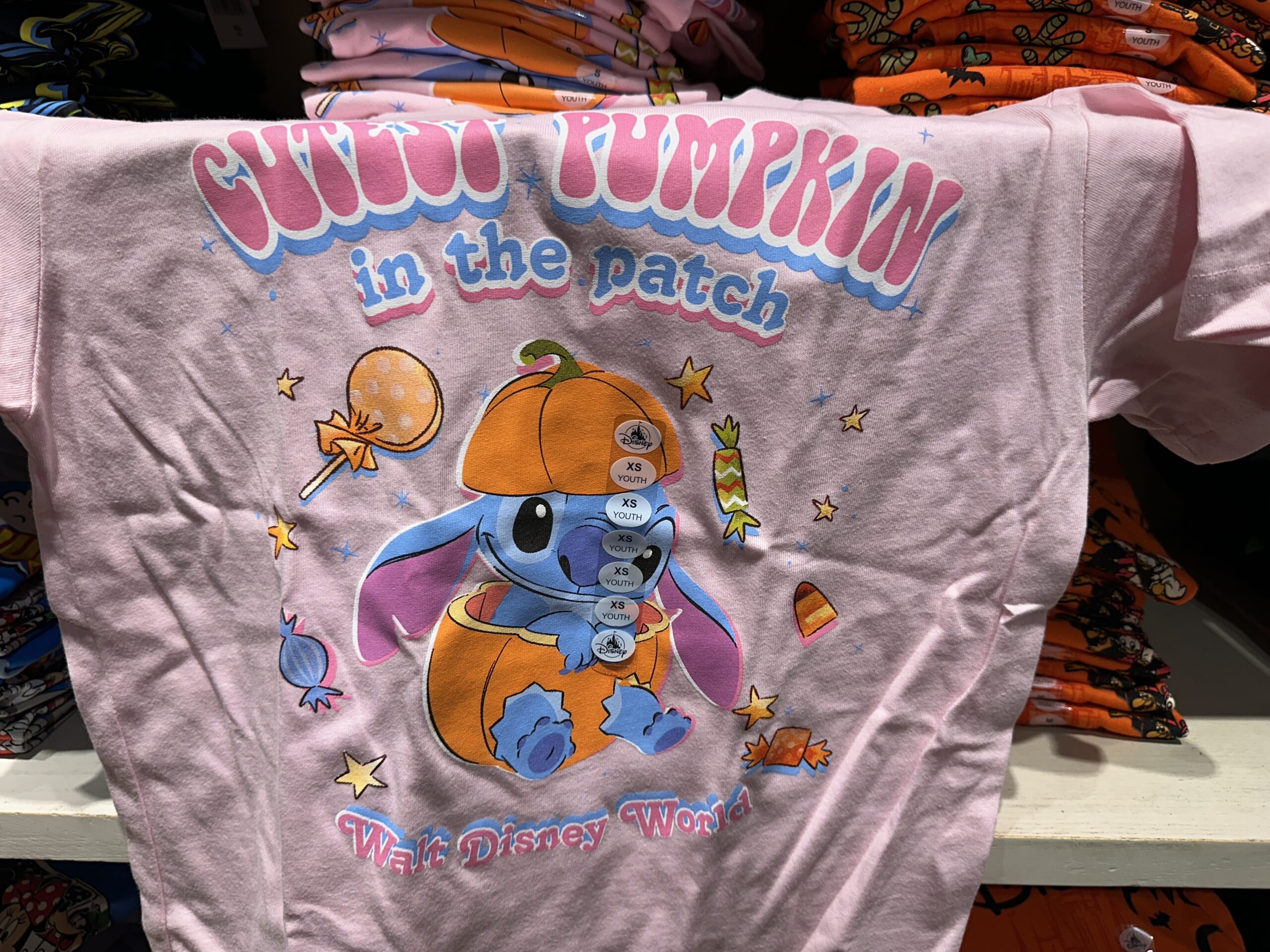 Stitch sits inside a pumpkin on this pink tee as Halloween candy surrounds him. "Cutest Pumpkin in the patch" is at the top in pink and blue text. "Walt Disney World" is at the bottom in the same style.