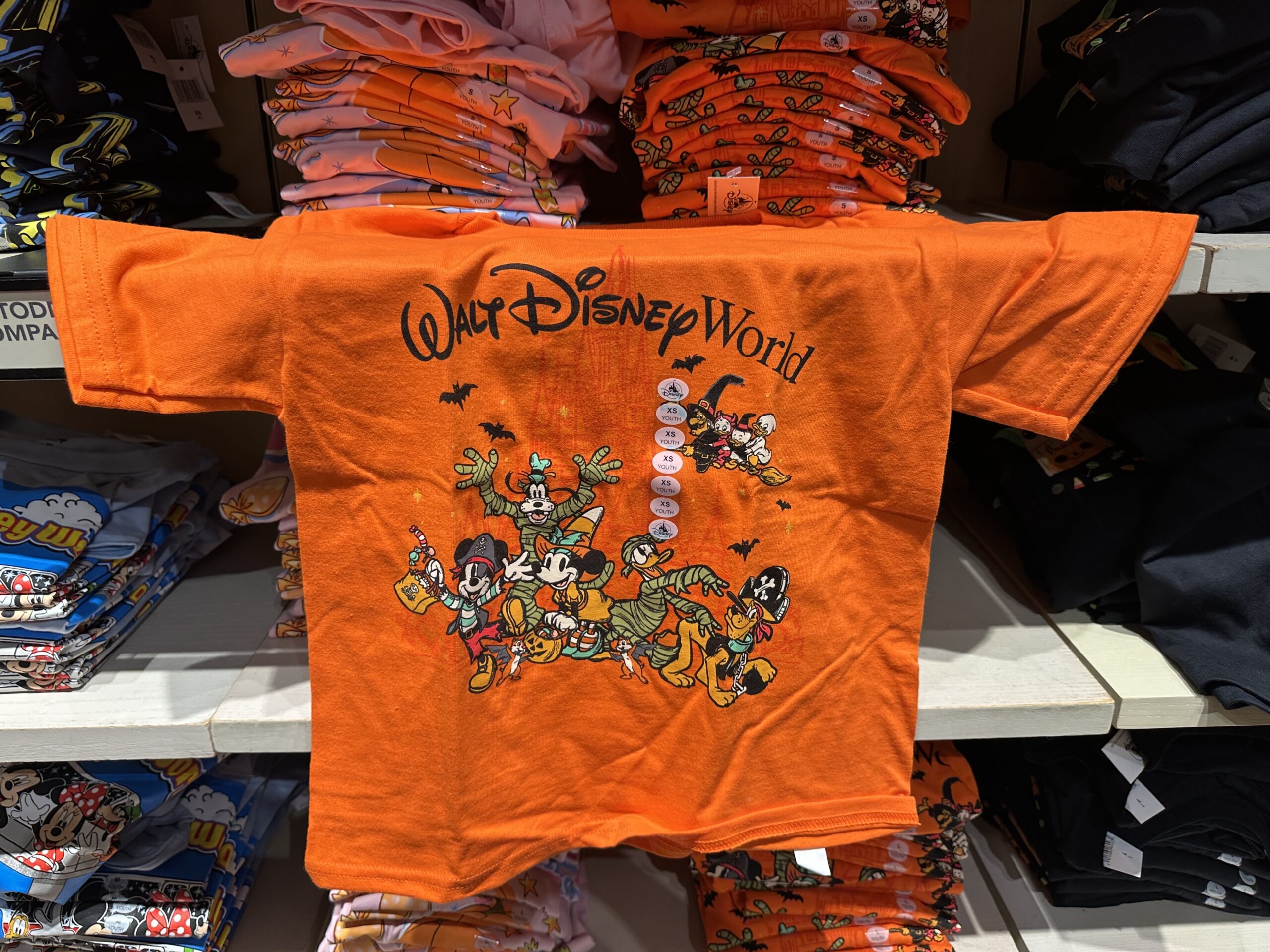 This orange tee has images of Mickey and his friends dressed in various costumes. Mickey is a pirate, Minnie is a witch, Goofy and Donald are both mummies, and Pluto is also a pirate. Behind them, bats fly and a witch carries Huey, Dewey, and Louie on a broom. The Walt Disney World wordmark is at the top in black.