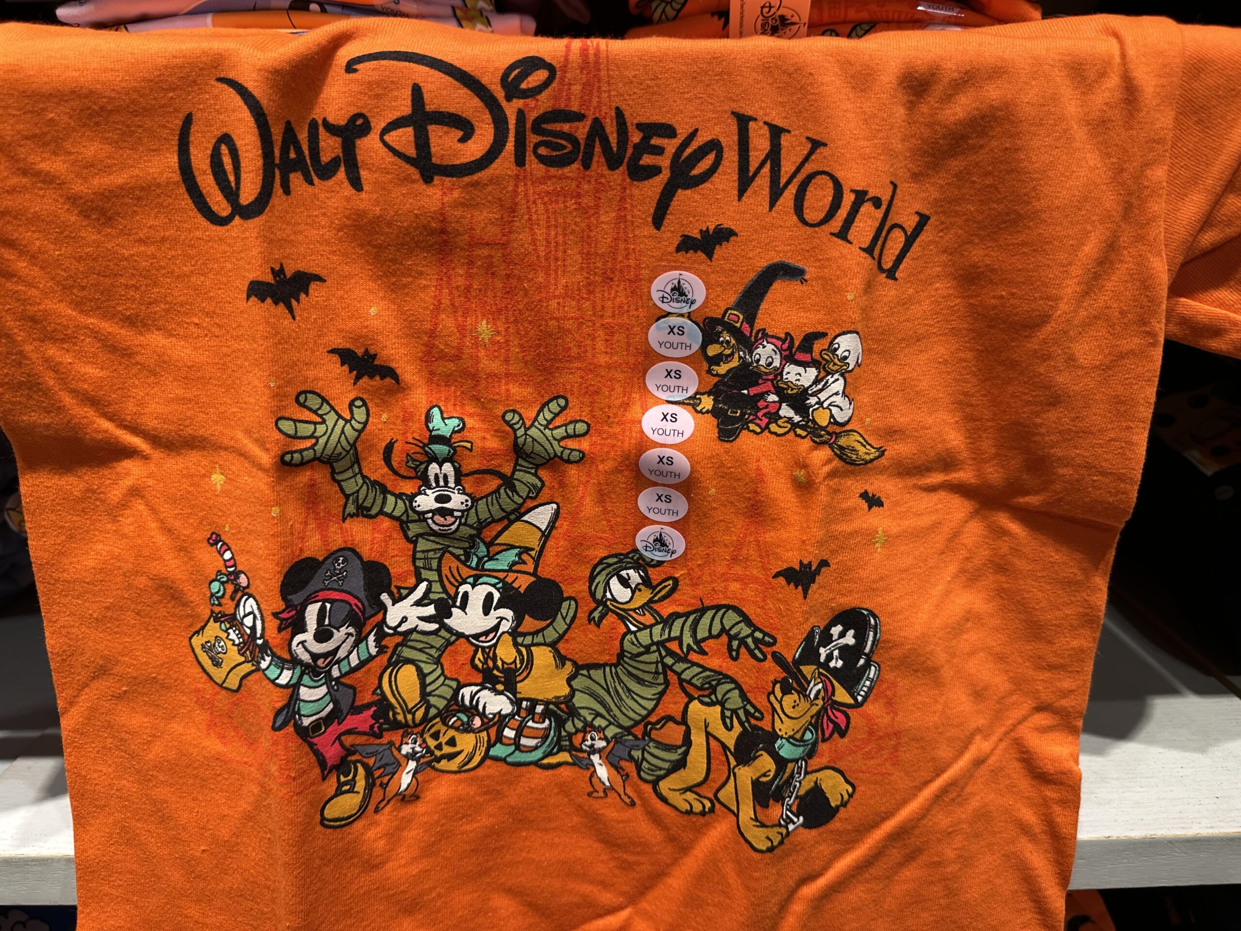 This orange tee has images of Mickey and his friends dressed in various costumes. Mickey is a pirate, Minnie is a witch, Goofy and Donald are both mummies, and Pluto is also a pirate. Behind them, bats fly and a witch carries Huey, Dewey, and Louie on a broom. The Walt Disney World wordmark is at the top in black.