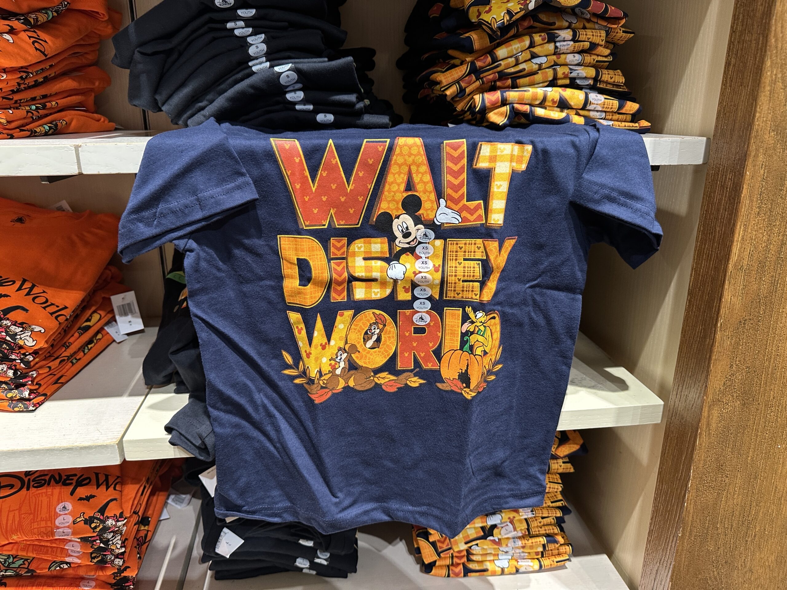 Mickey, Chip ‘n’ Dale, and Pluto are celebrating autumn on this autumnal shirt. The T-shirt is navy blue with “Walt Disney World” in patterned fall colors across the front. Acorns and autumn leaves are scattered beneath the wording, and a Fantasyland Castle Jack-o’-lantern is on the far right.