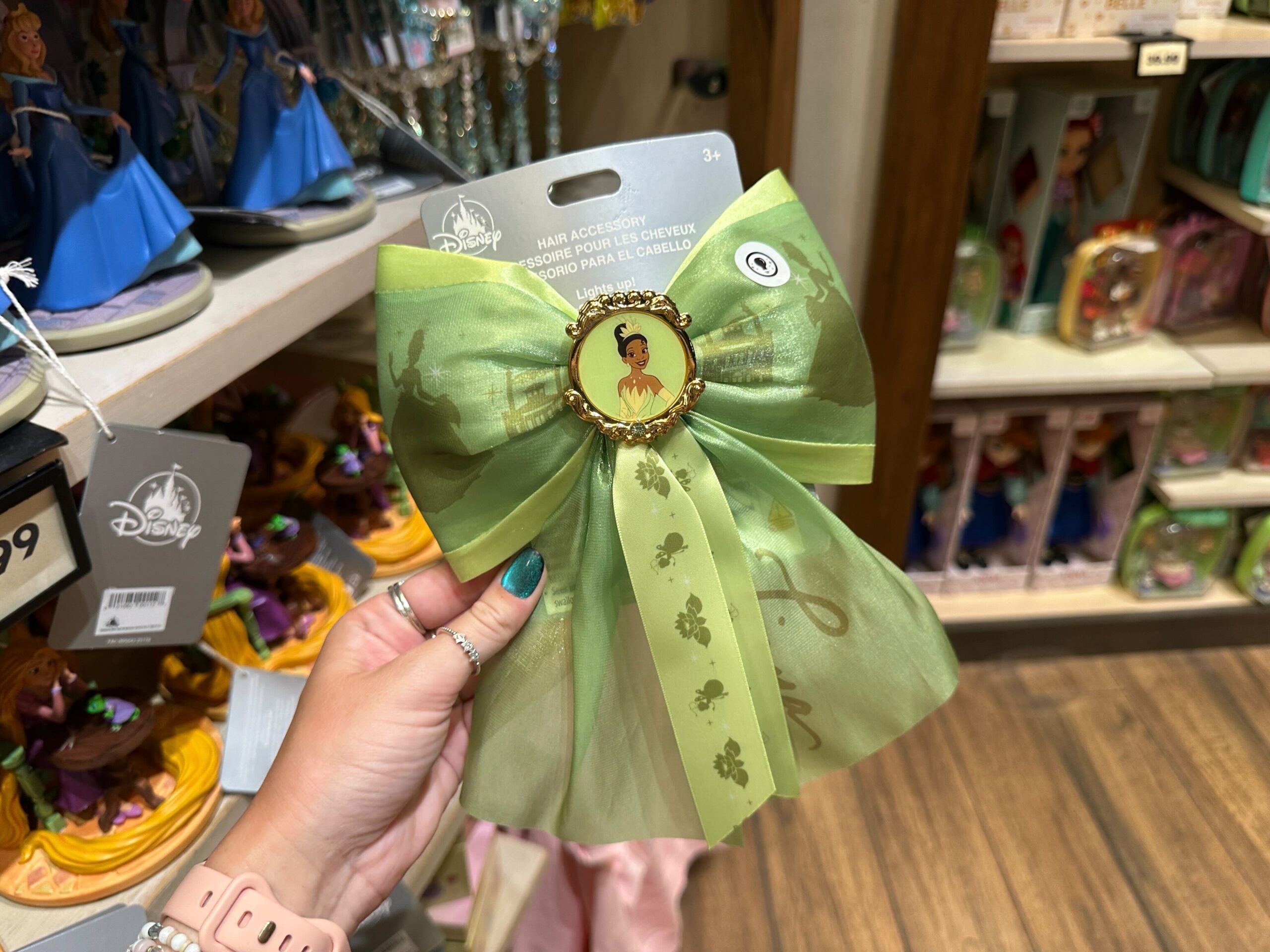 Princess Tiana has her own light and dark green hair bow, complete with her portrait in the middle and silhouettes of her standing by her restaurant on the ribbon. "Tiana" is written in cursive on one bow tail, while other bow tails have a repeating pattern of water lilies and Ray the firefly.