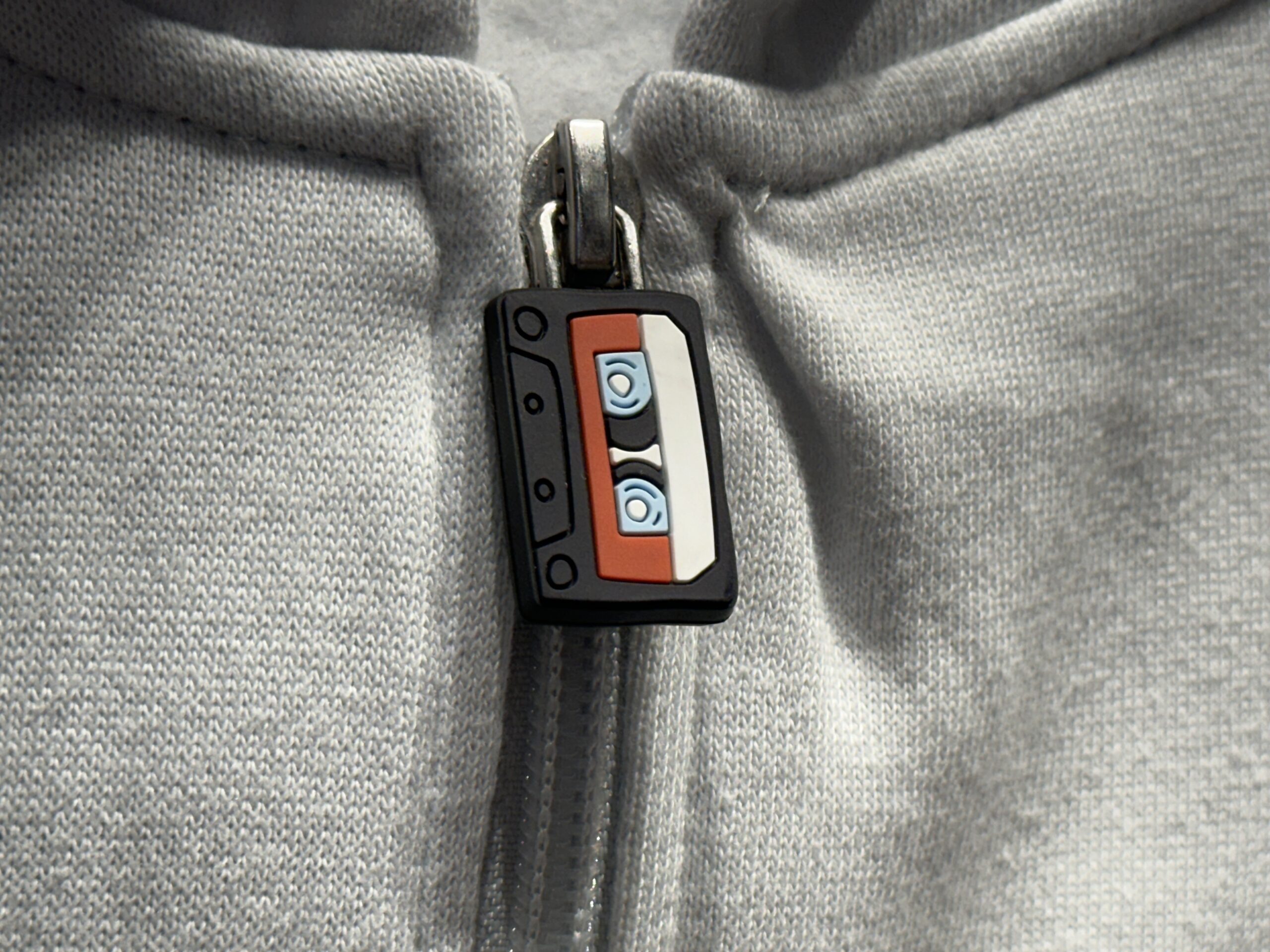 This white hoodie has kangaroo pockets and zips up with a silicone cassette tape zipper pull. The Guardians of the Galaxy: Cosmic Rewind wordmark is embroidered on the left chest.