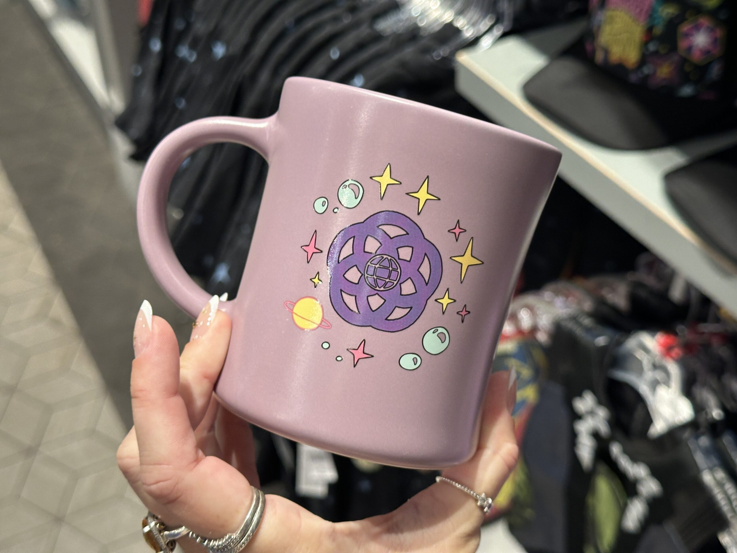 The mug is the same lilac color as the sweatshirt with the purple and pink "Guardians of the Galaxy: Cosmic Rewind" art on one side. A purple version of the EPCOT logo is on the reverse with a ring of planets, bubbles, and stars around it.