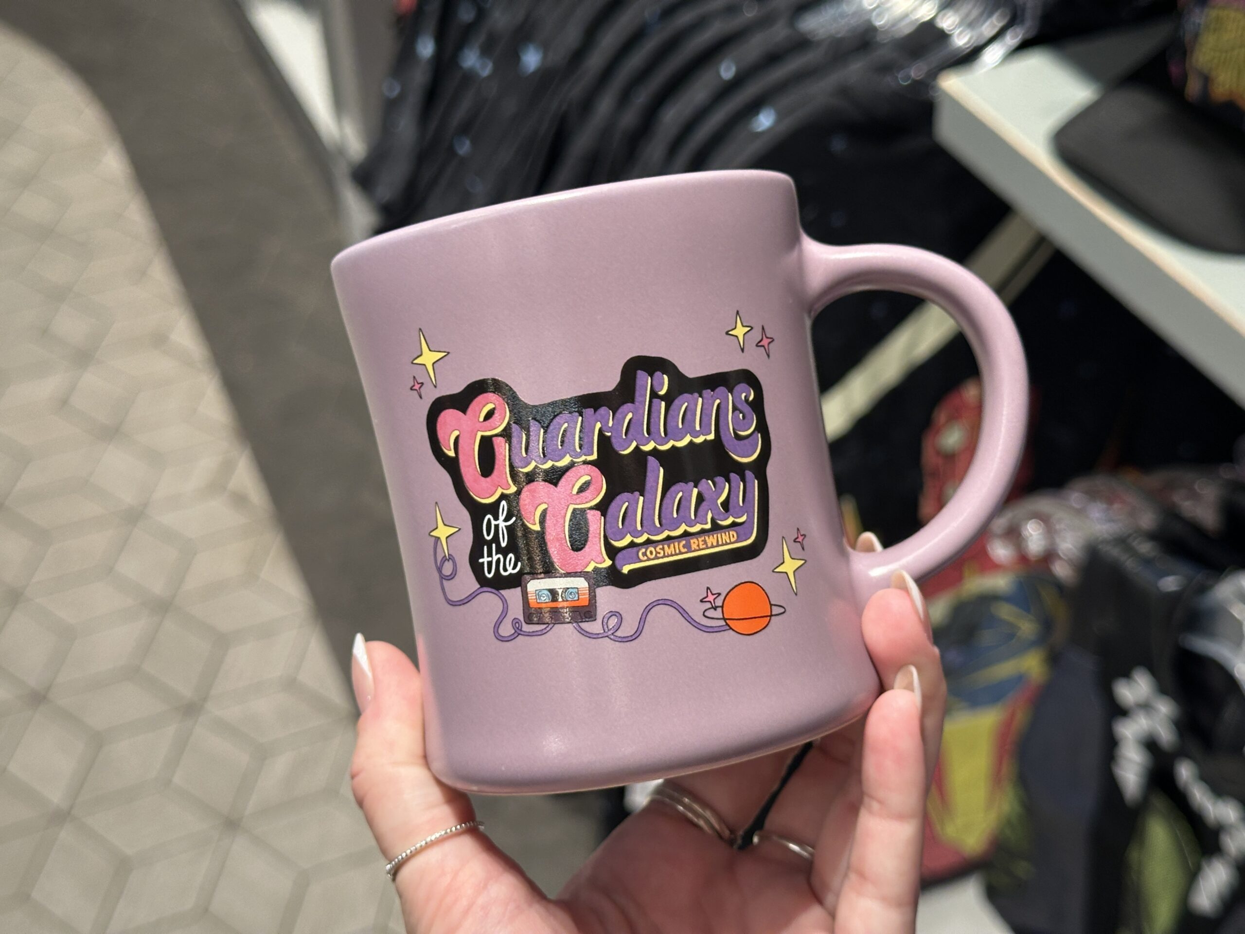 The mug is the same lilac color as the sweatshirt with the purple and pink "Guardians of the Galaxy: Cosmic Rewind" art on one side. A purple version of the EPCOT logo is on the reverse with a ring of planets, bubbles, and stars around it.