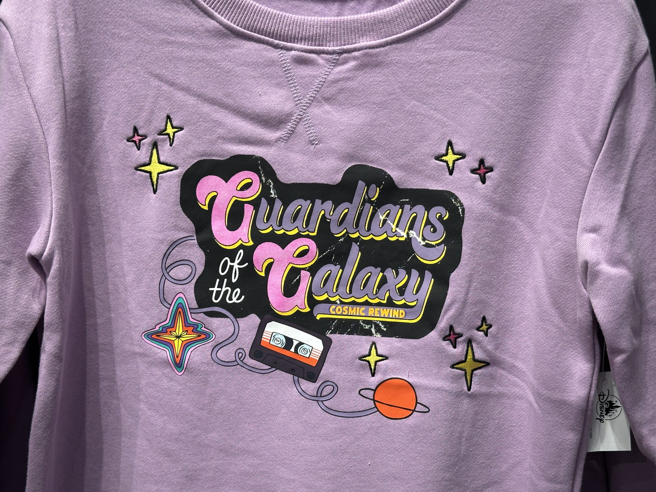 "Guardians of the Galaxy: Cosmic Rewind" is on the chest of this lilac sweatshirt in purple, pink, white, and yellow against a black background. Around the text are embroidered stars, the Nova Corps logo, a planet, and a cassette with its tape unwinding.