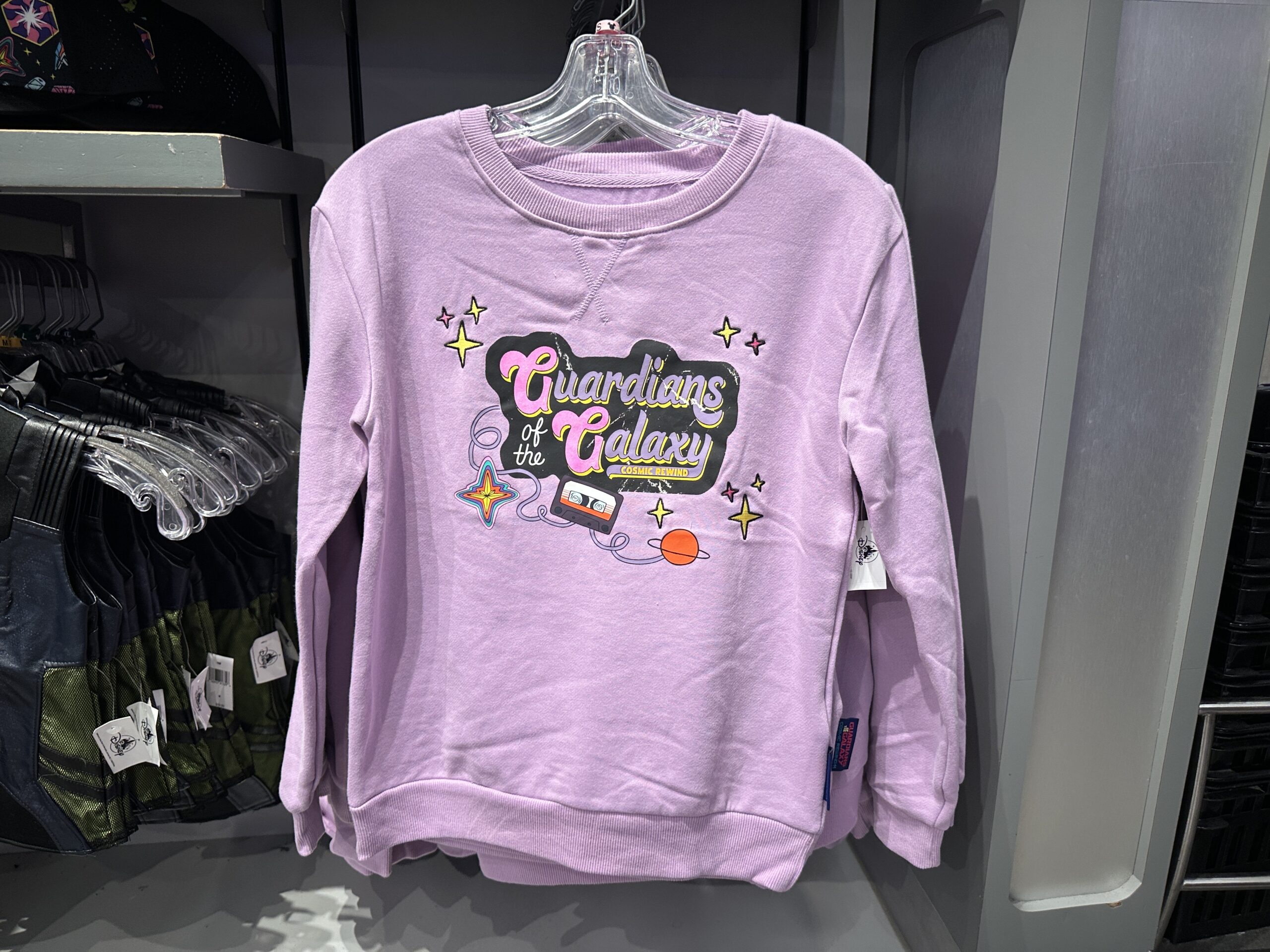 "Guardians of the Galaxy: Cosmic Rewind" is on the chest of this lilac sweatshirt in purple, pink, white, and yellow against a black background. Around the text are embroidered stars, the Nova Corps logo, a planet, and a cassette with its tape unwinding.