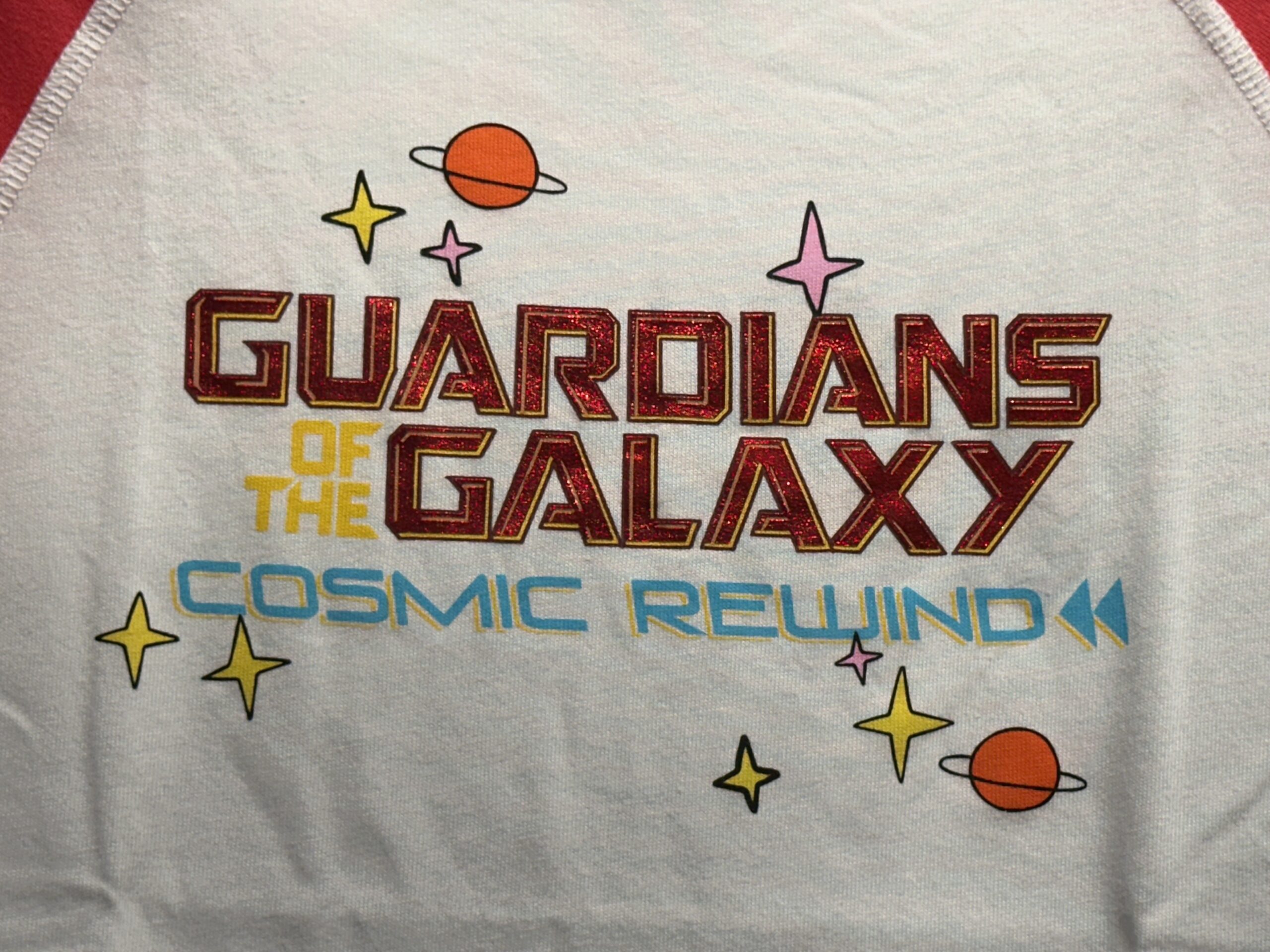 This is a white tee with red short sleeves and a red neckline. "Guardians of the Galaxy: Cosmic Rewind" is on the chest. "Guardians" and "Galaxy" are in shiny red lettering, while "of the" is yellow and "Cosmic Rewind" is blue. Around the text are minimalist planets and stars.