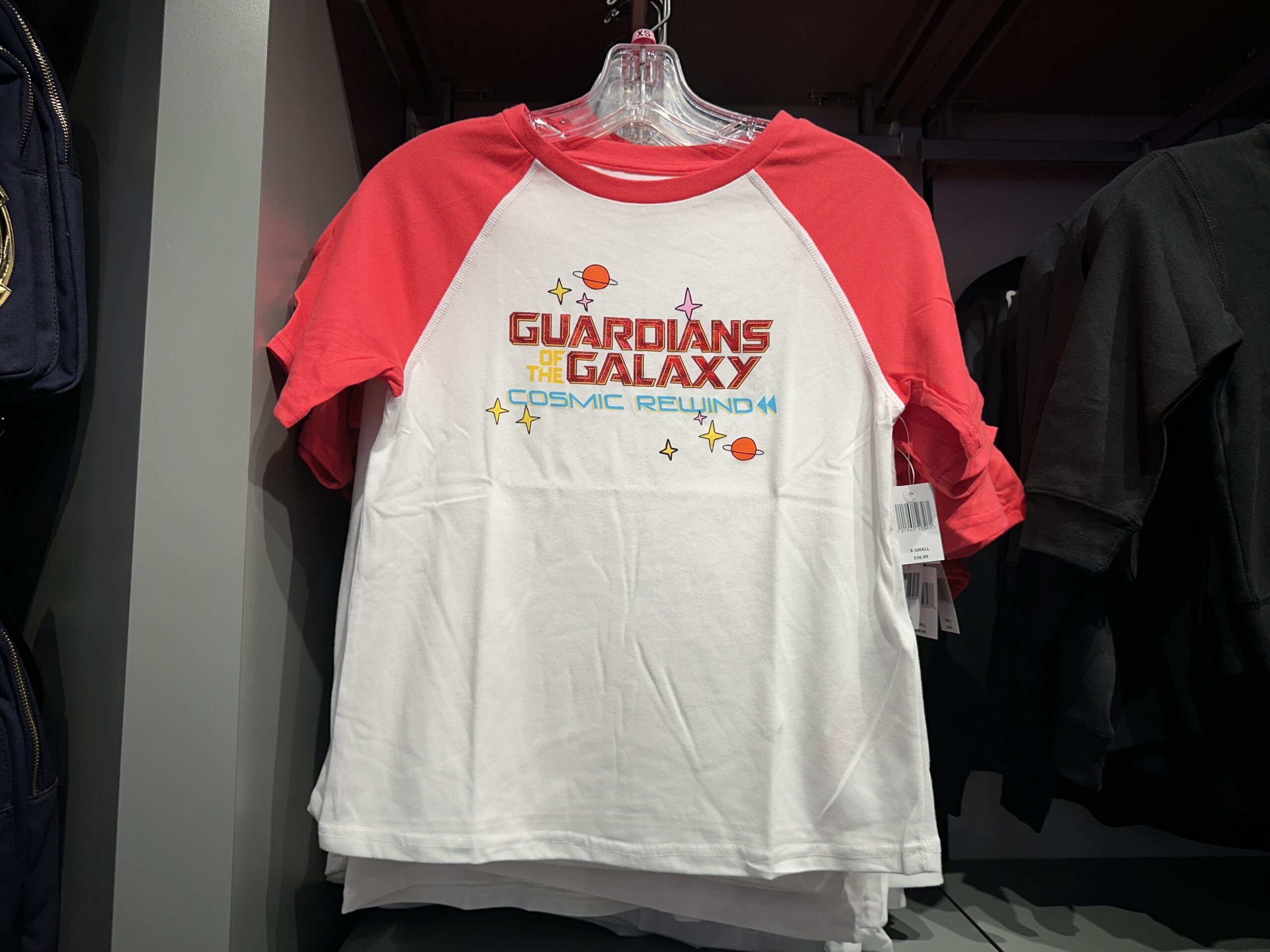 This is a white tee with red short sleeves and a red neckline. "Guardians of the Galaxy: Cosmic Rewind" is on the chest. "Guardians" and "Galaxy" are in shiny red lettering, while "of the" is yellow and "Cosmic Rewind" is blue. Around the text are minimalist planets and stars.