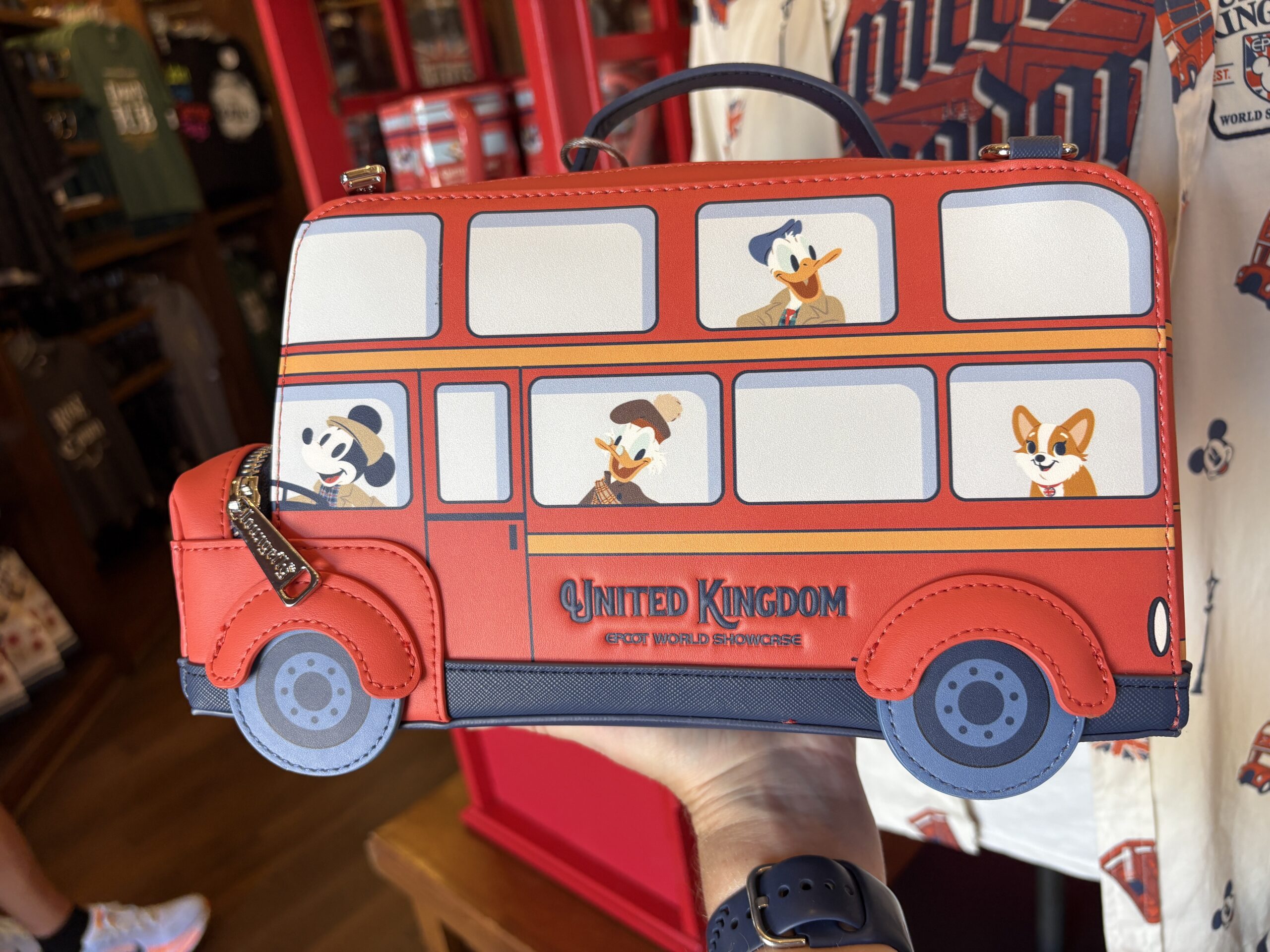 The faux leather bag is shaped like a red double-decker bus with a navy blue base and wheels. Printed on the sides are images of Disney characters (and a corgi) in the windows of the bus. Mickey is behind the wheel.