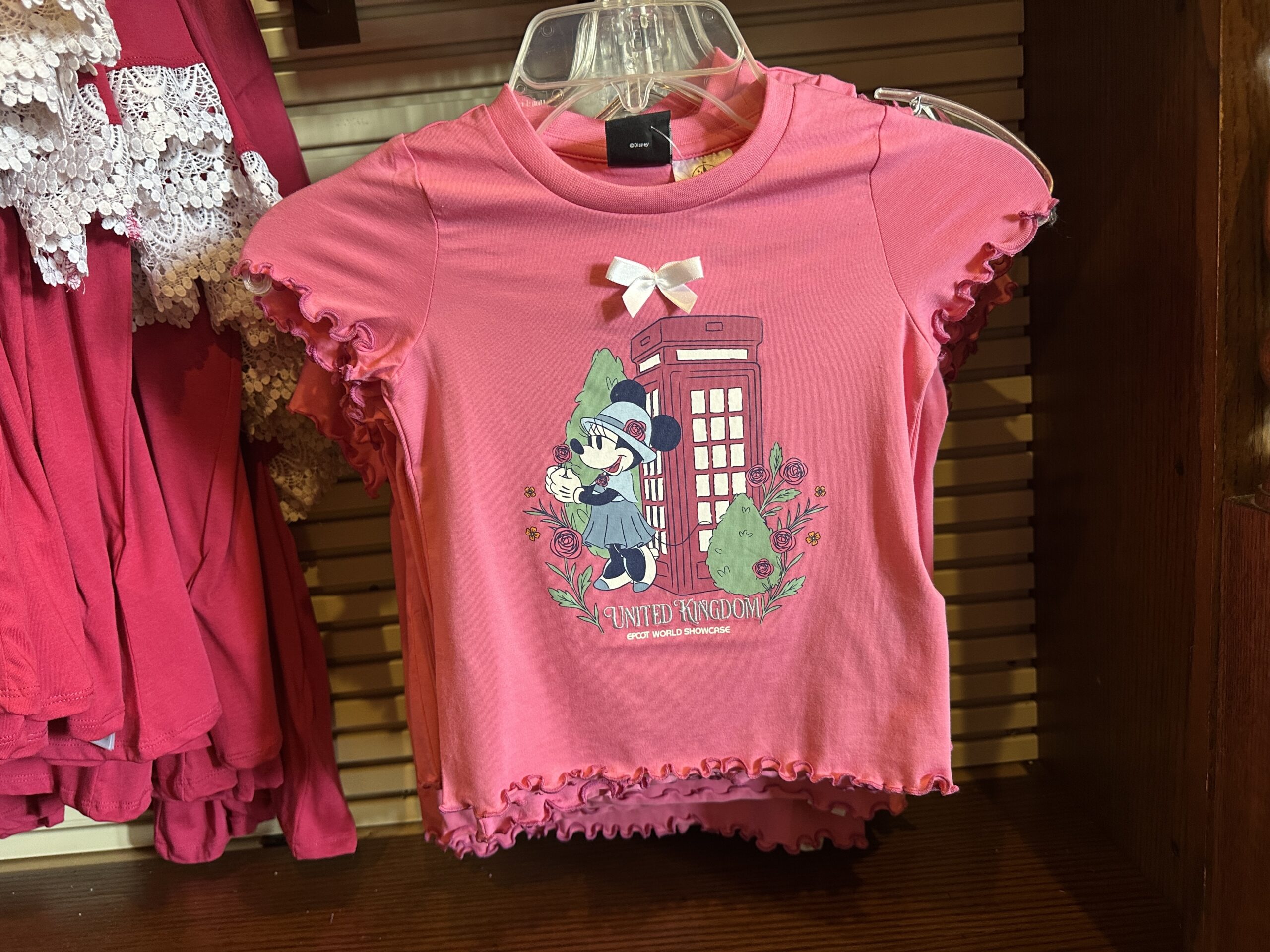 Minnie is also pictured on the youth tee in her blue U.K. outfit, alongside roses and a red telephone booth. A white satin bow is attached above the art. The tee has ruffled edges.