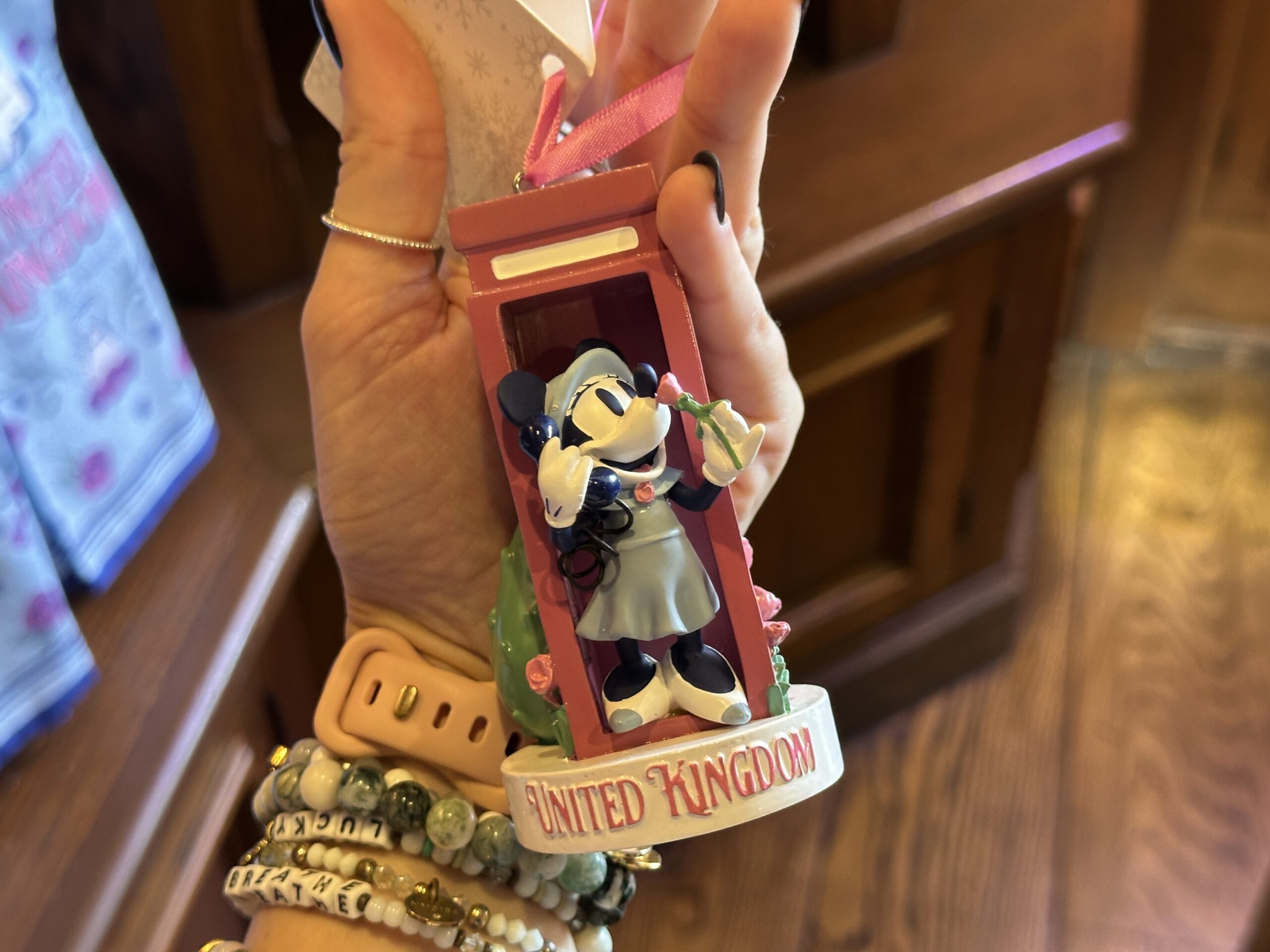 This sculpted ornament depicts Minnie talking on the phone, leaning out of a red booth with a rose to her nose. The circular base features "United Kingdom" on the front.