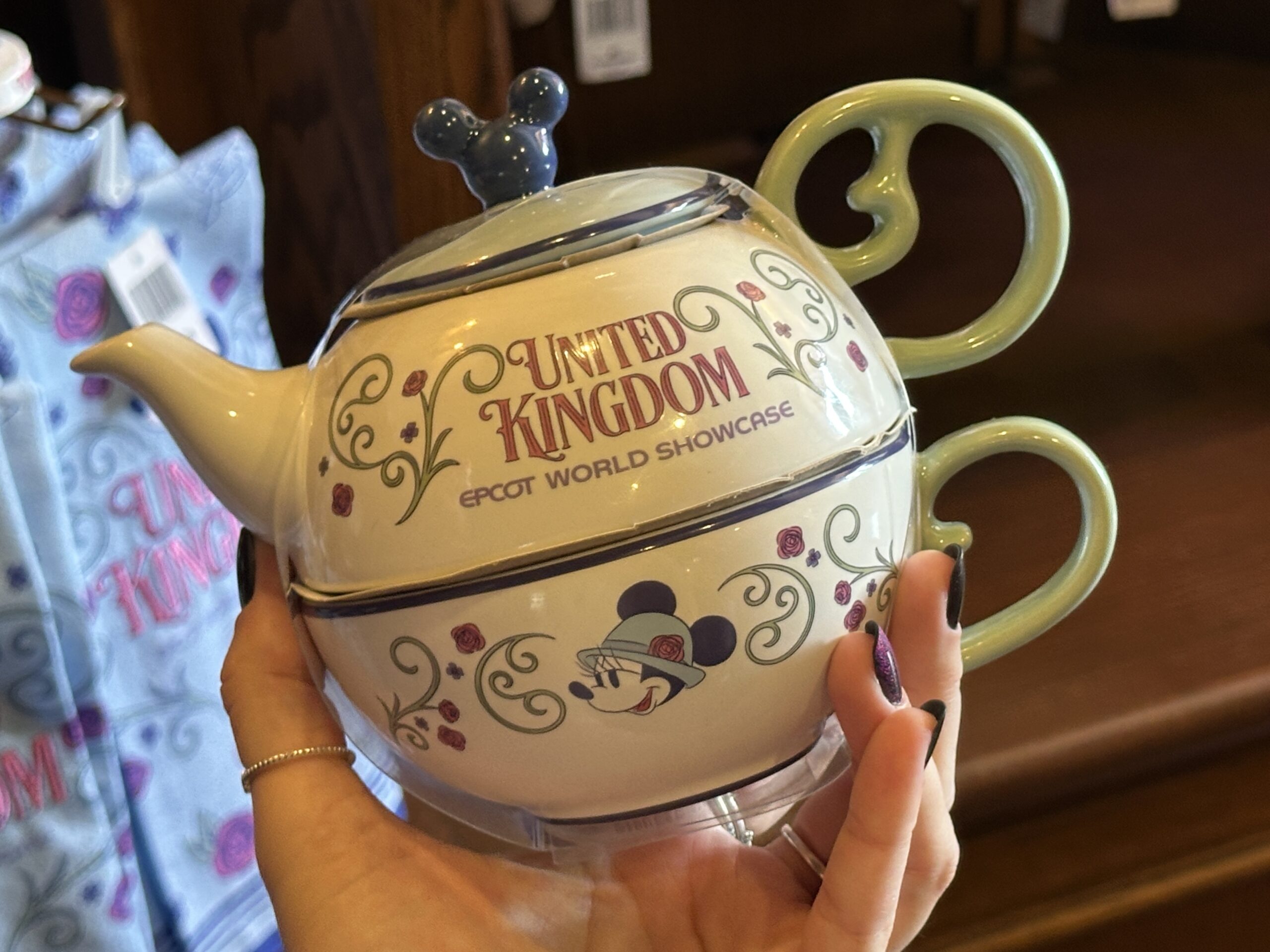 This set includes a small tea pot and tea cup, which stack together. They are white stoneware with green vine-like handles. The pot, which sits on top of the cup, reads "United Kingdom EPCOT World Showcase." It has a blue lid with a Mickey-shaped topper.