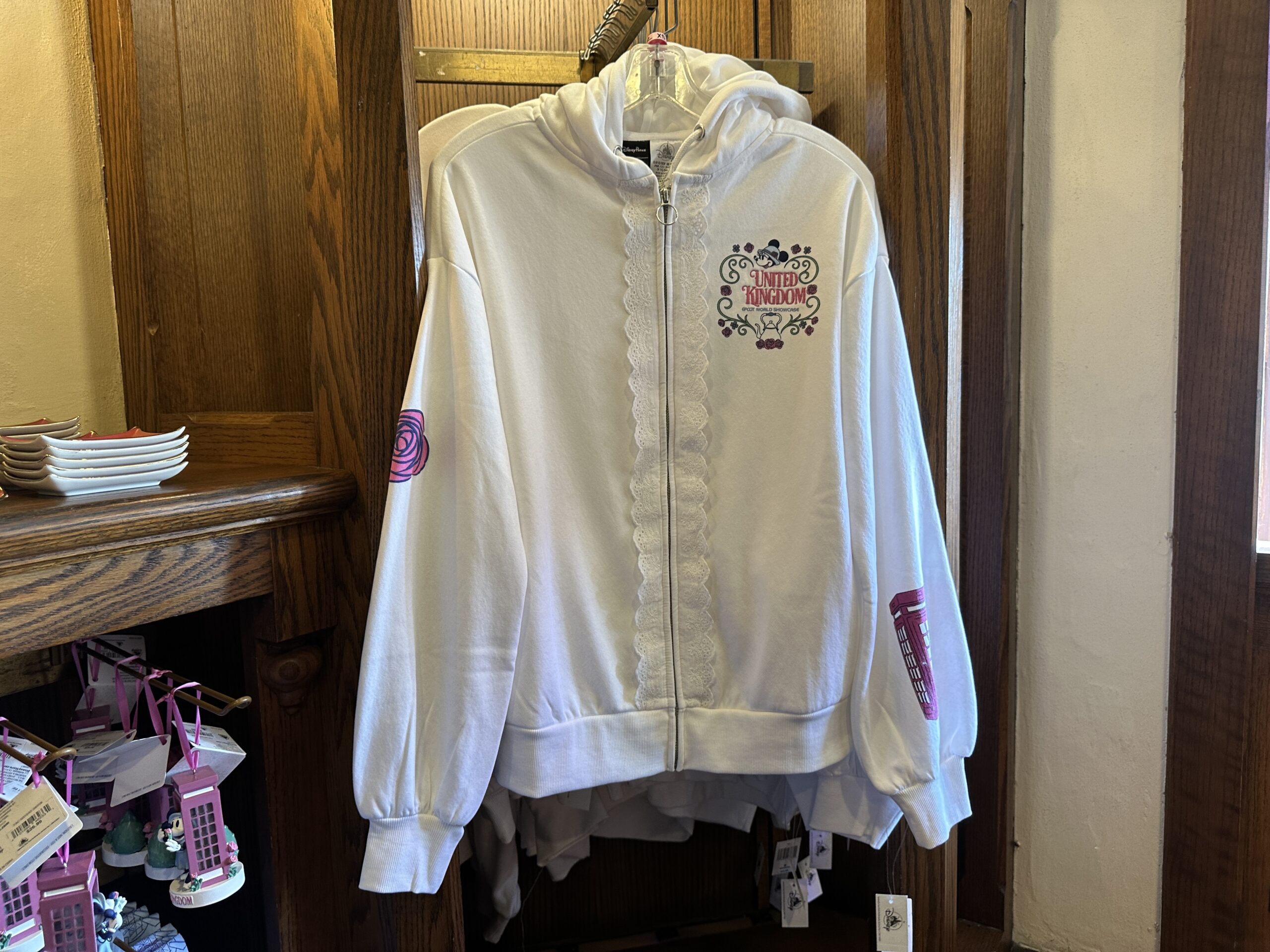 Lace lines the zipper on the front of this white hoodie. It has bishop sleeves. "United Kingdom" is embroidered on the left chest with an image of Minnie, a tea pot, and roses.