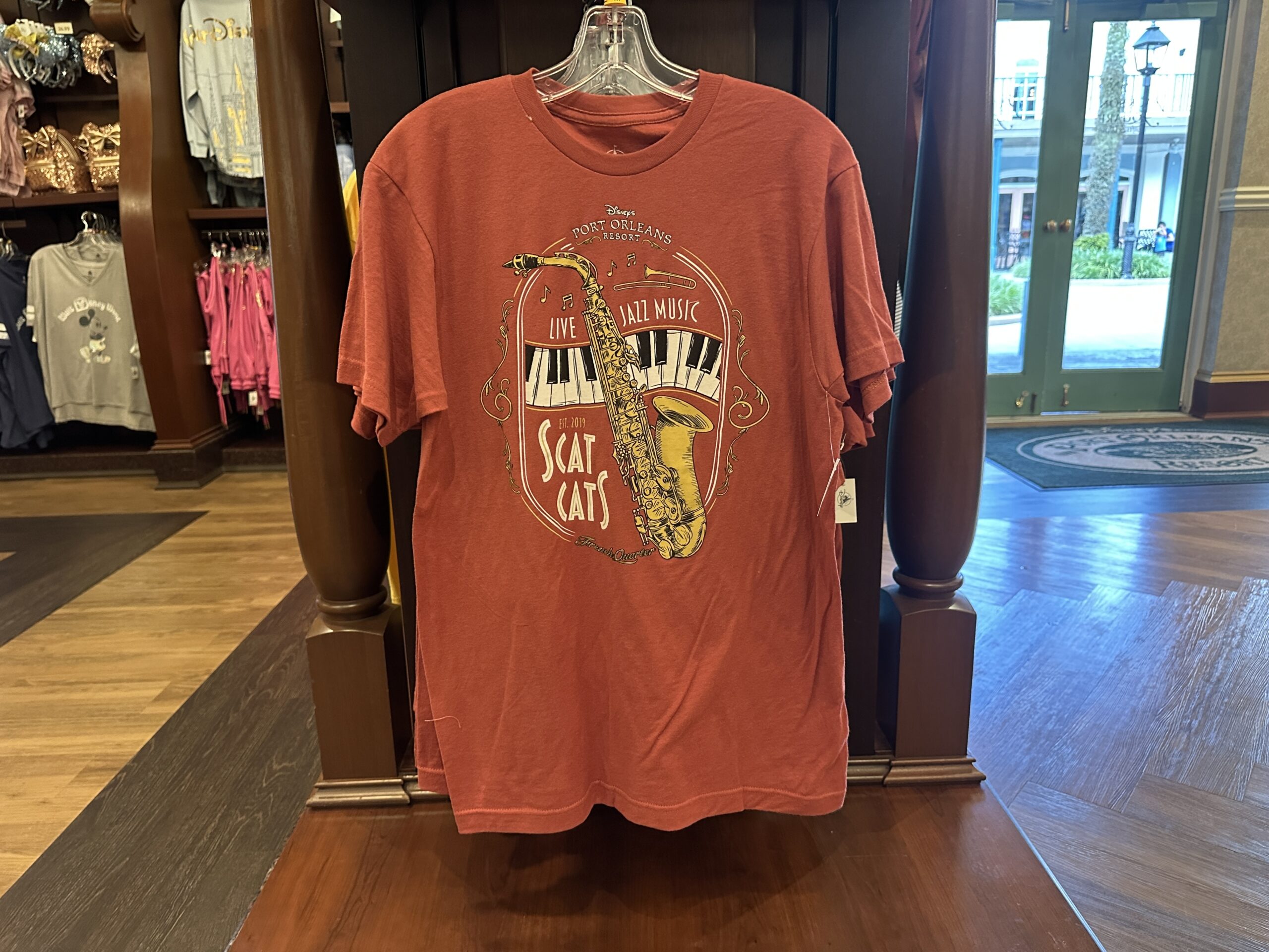 This red tee has artwork of a saxophone, curved piano keys, a trumpet, and music notes. Disney's Port Orleans Resort is at the top. "Live Jazz Music" is above the keys.
