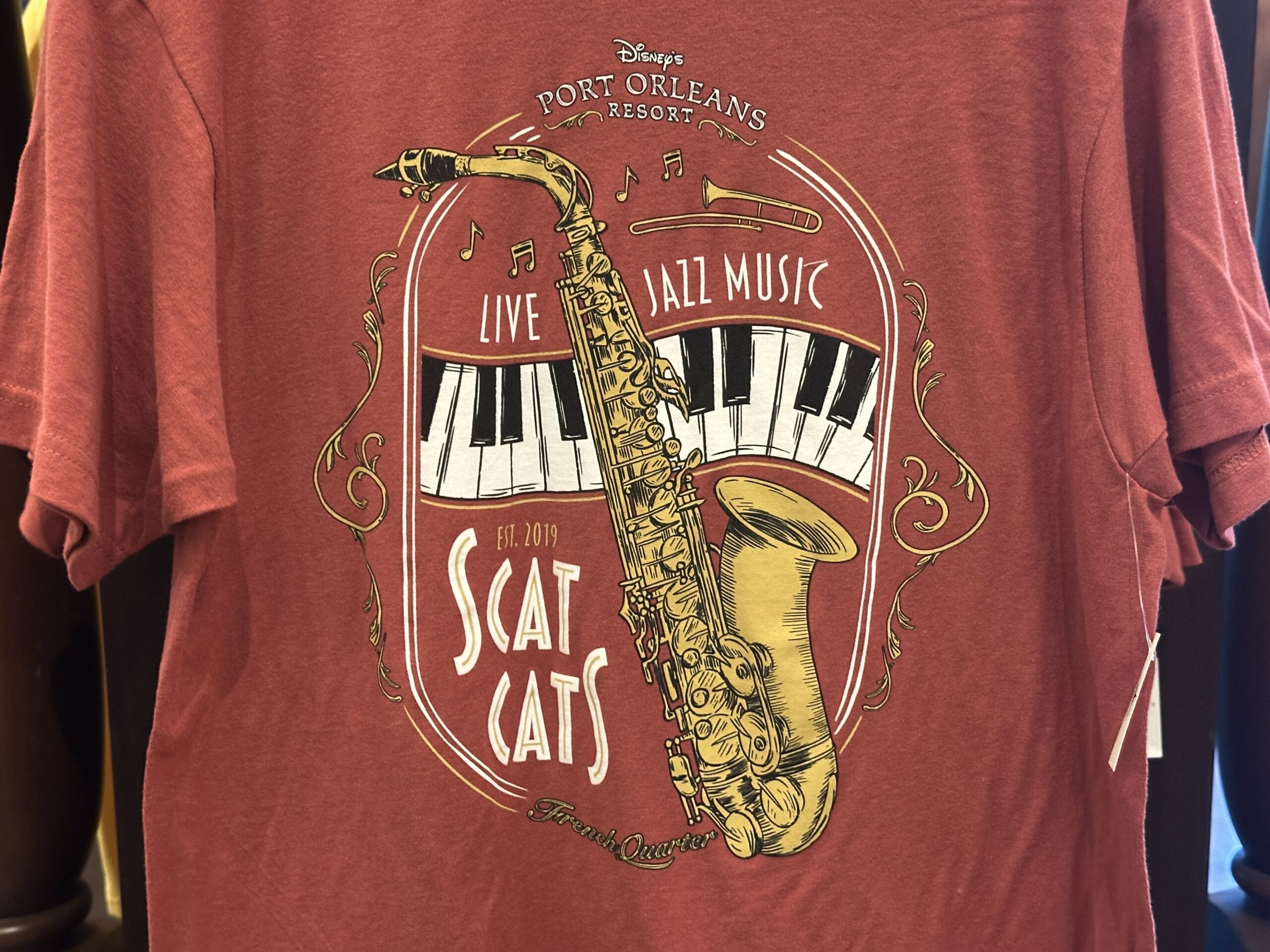 This red tee has artwork of a saxophone, curved piano keys, a trumpet, and music notes. Disney's Port Orleans Resort is at the top. "Live Jazz Music" is above the keys.